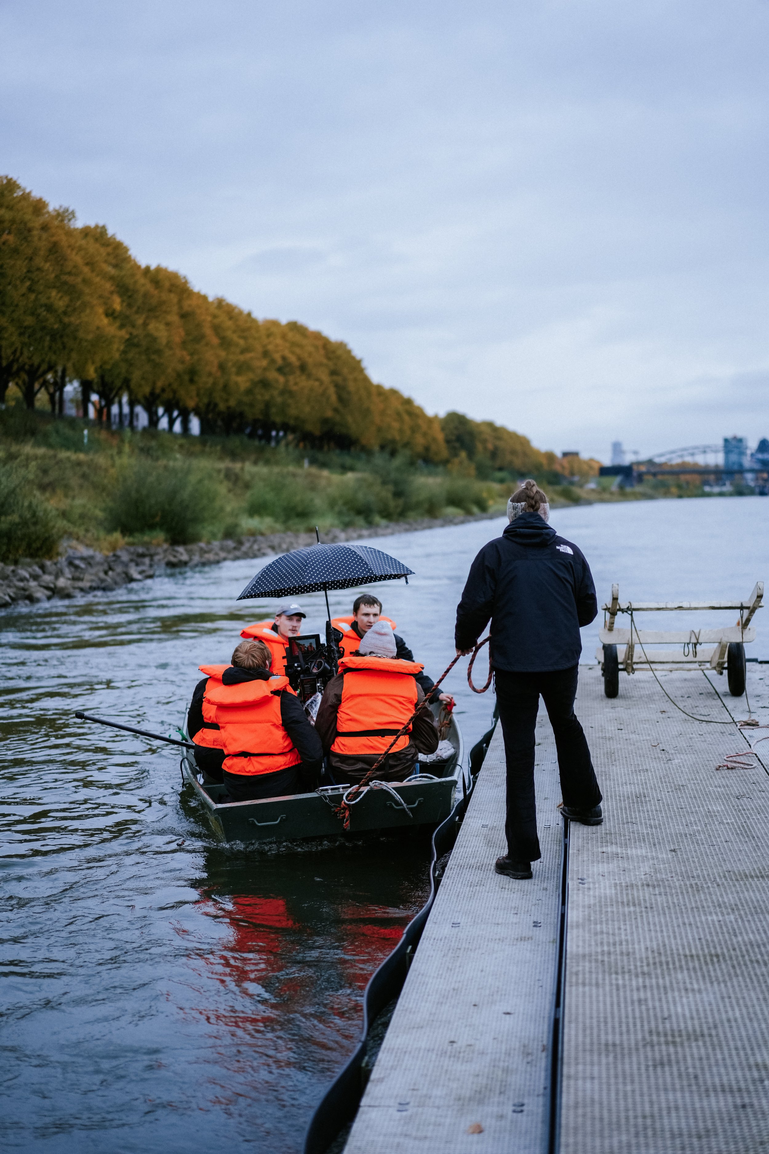 Menschen in einem Boot auf einem Fluss, geschützt durch orange Rettungswesten, während eine Person am Steg das Boot festmacht, unter einem Regenschirm, bei bewölktem Himmel.
