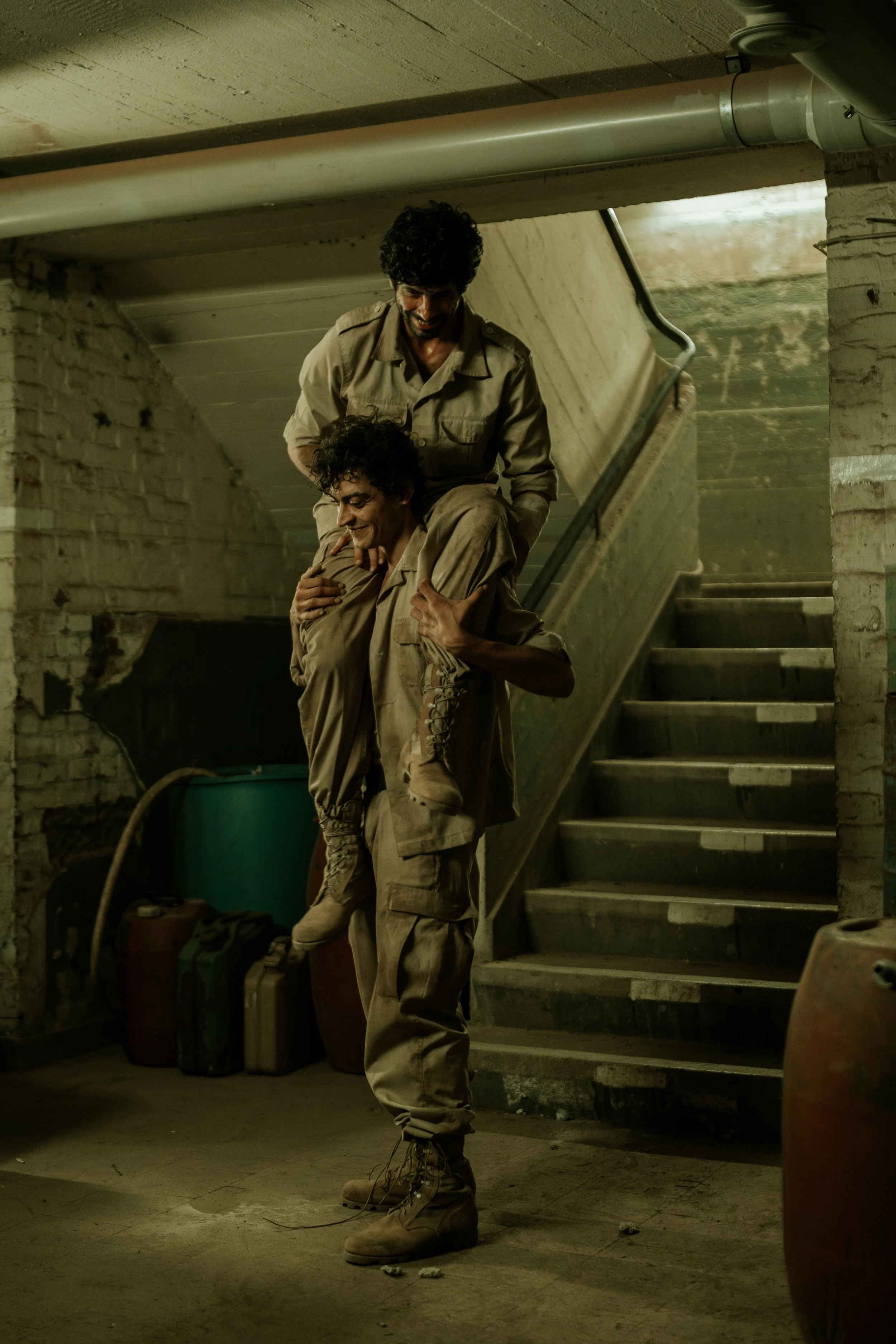 Zwei Männer in Kampfuniformen in einem Keller, einer trägt den anderen auf den Schultern, beide lachen.