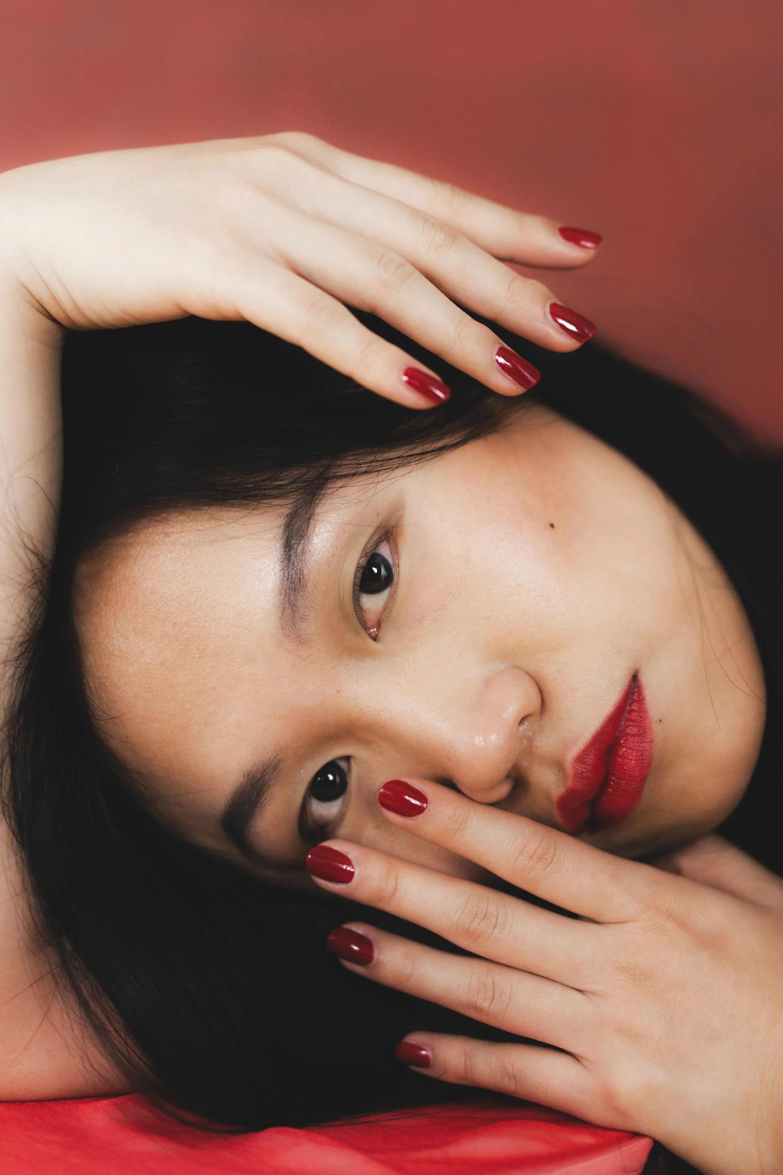 Nahaufnahme eines asiatisches Mädchens mit roten Lippen und rotem Nagellack, das auf einer Oberfläche liegt und den Blick in die Kamera gerichtet hat.