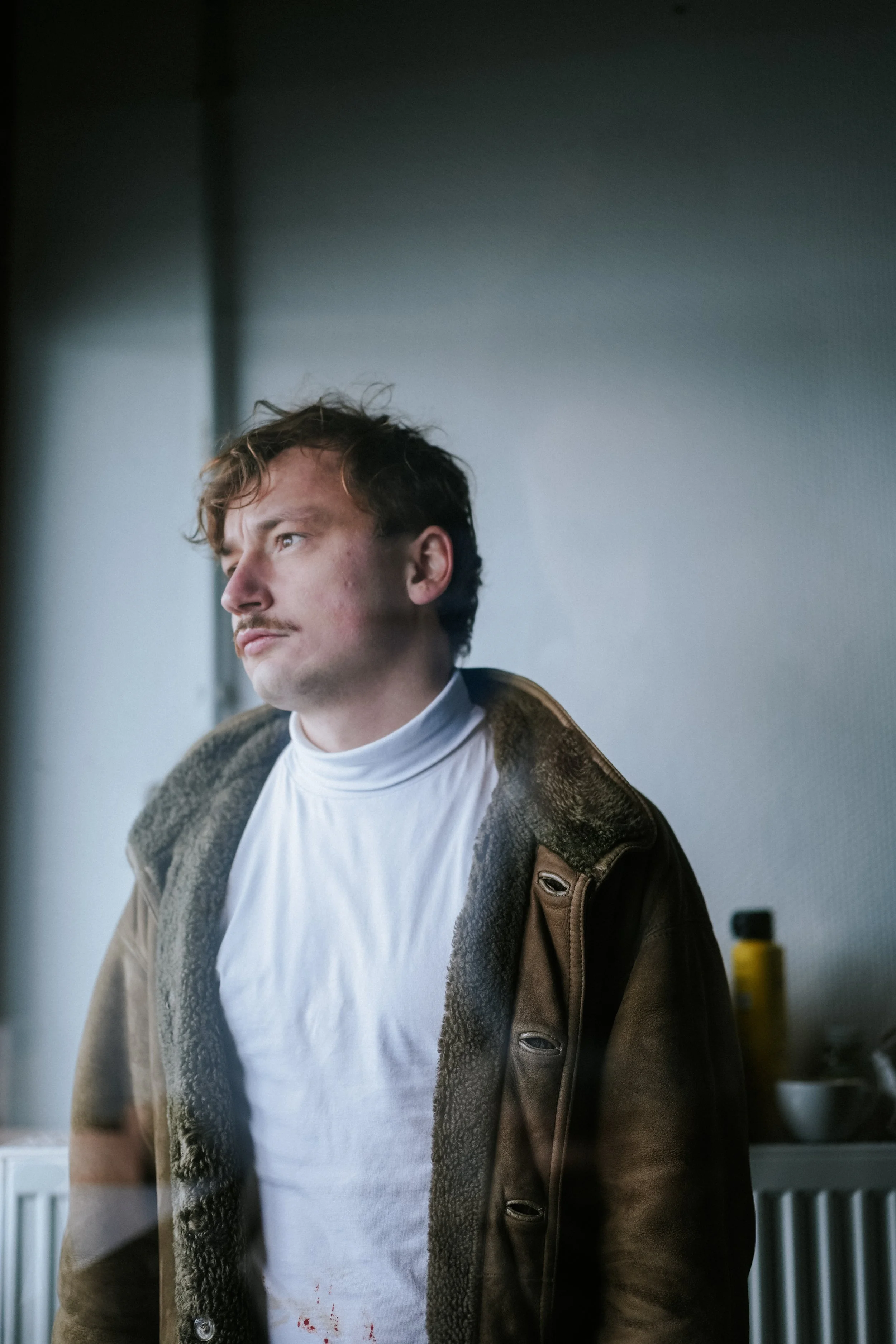 Junger Mann mit einem Schnurrbart, trägt einen weißen Rollkragenpulli und eine braune Jacke, steht vor einer Wand, blickt nach links.