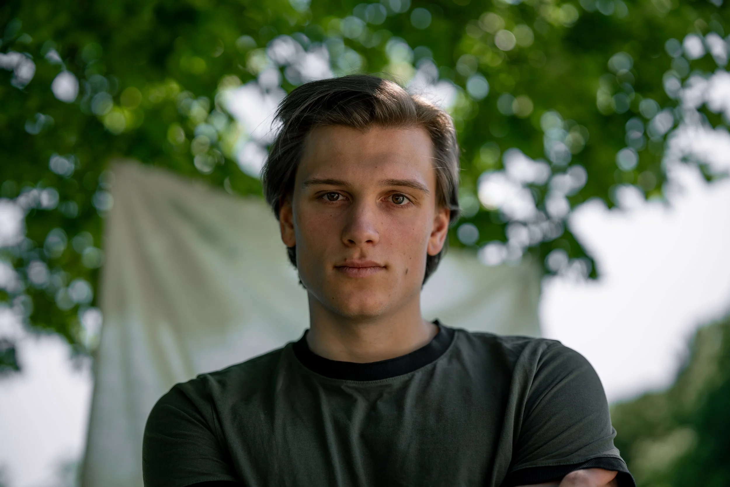 Junger Mann mit dunklem Hemd steht vor einem Baum mit grünen Blättern, im Hintergrund eine gebleichte weiße Tuch. Nahaufnahme, Blick direkt in die Kamera.