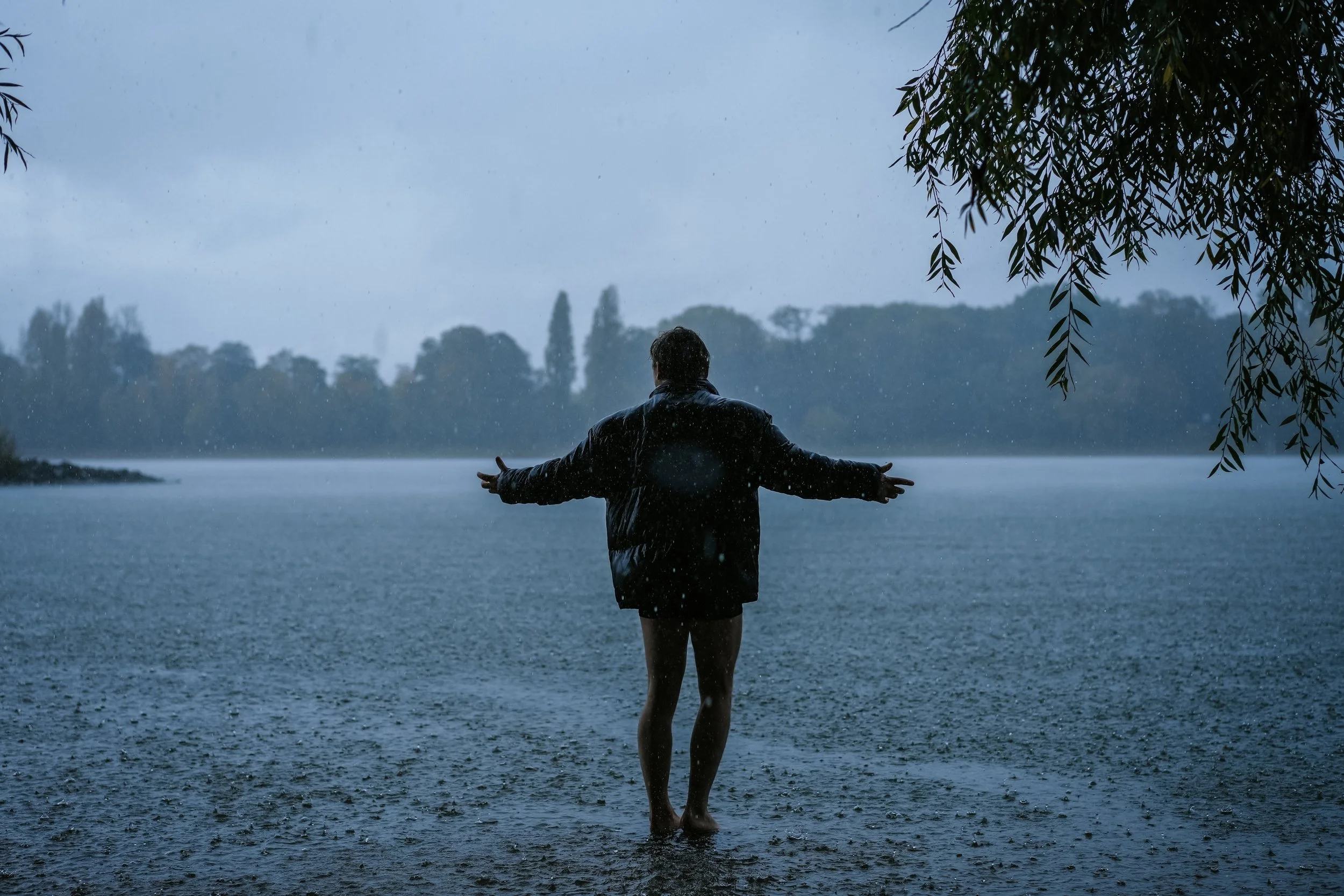 Eine Person steht barfuß im Regen an einem See mit offenen Armen, im Hintergrund bewölkter Himmel und Bäume, es ist regnerisch.