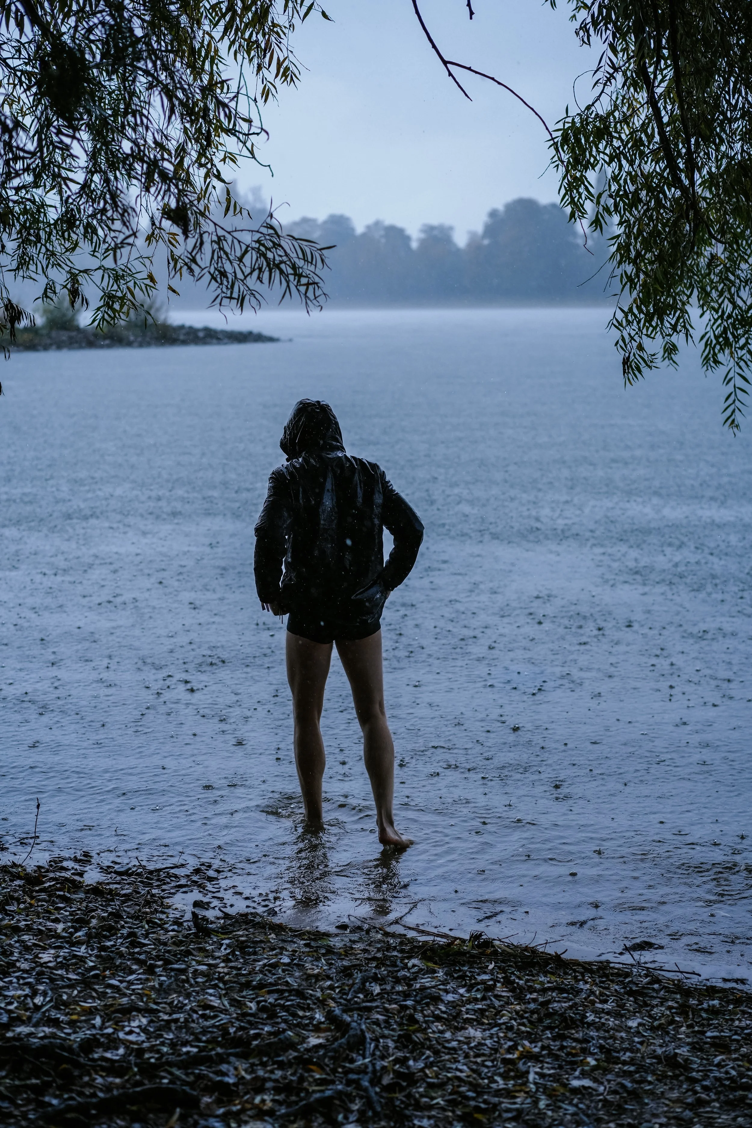 Person steht barfuß am Ufer eines Gewässers bei Regen, trägt eine Jacke, im Hintergrund bewölkter Himmel und Bäume.