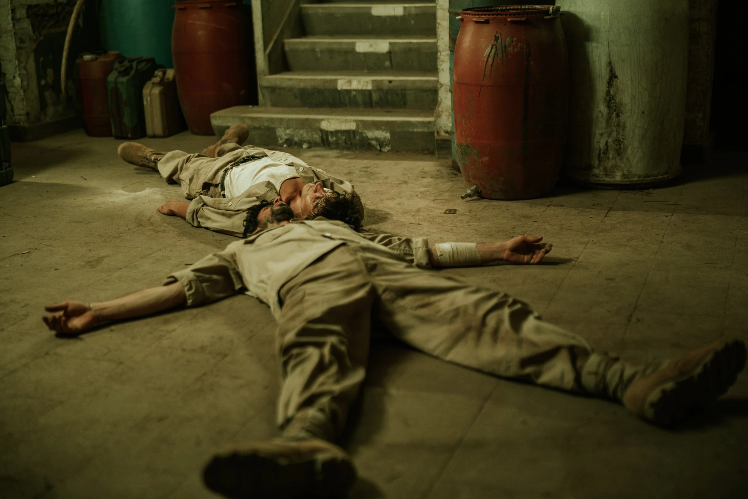 Zwei Männer liegen bewusstlos auf dem Boden in einem dunklen Raum mit gefässern und Treppen. Einer trägt eine Bandage am Arm.