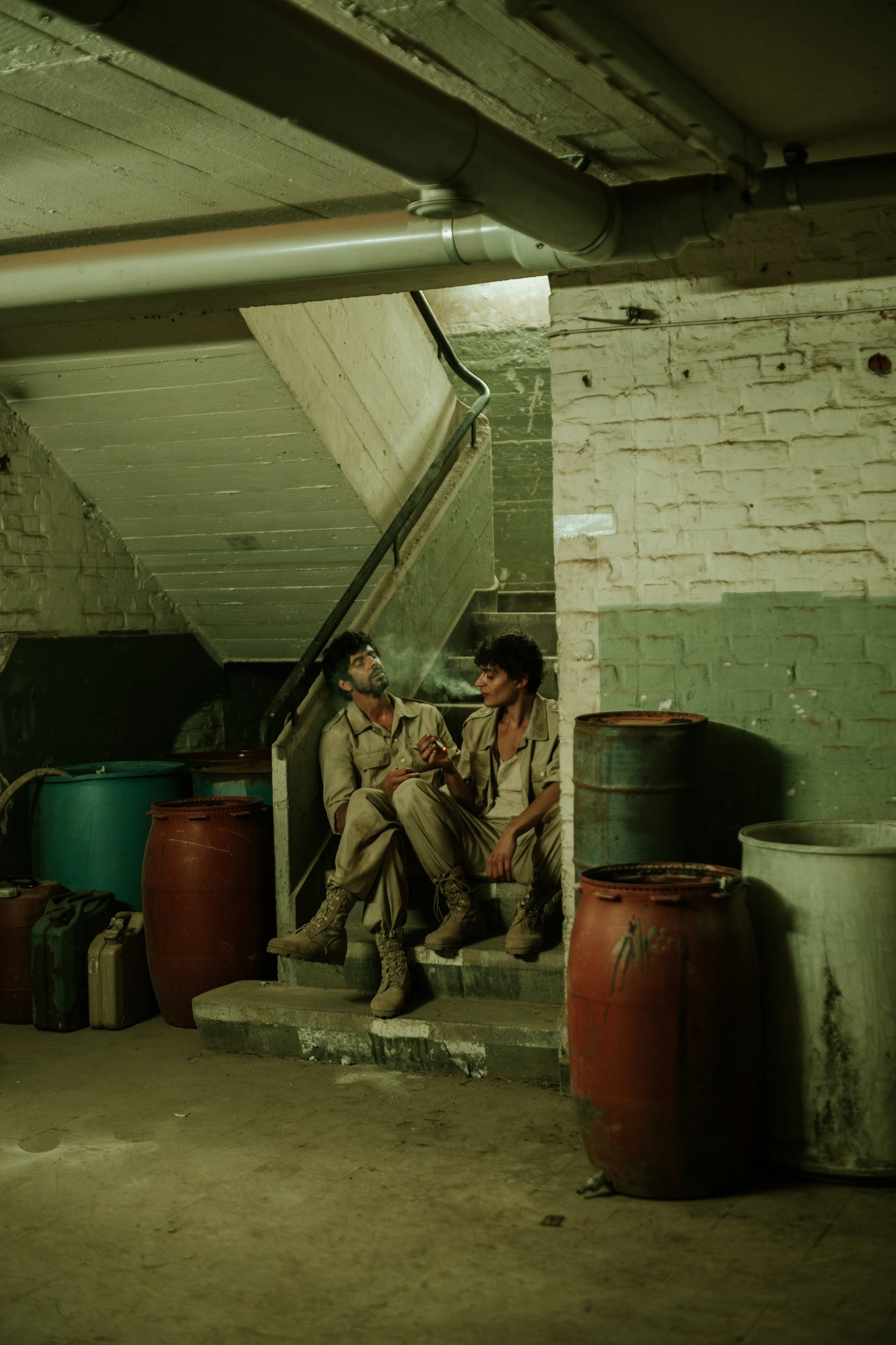 Zwei Menschen in Kriegskleidung sitzen auf einer Treppe in einer grauen, abgenutzten Umgebung mit Big Packs und Fässern um sie herum, bei schwachem Licht.