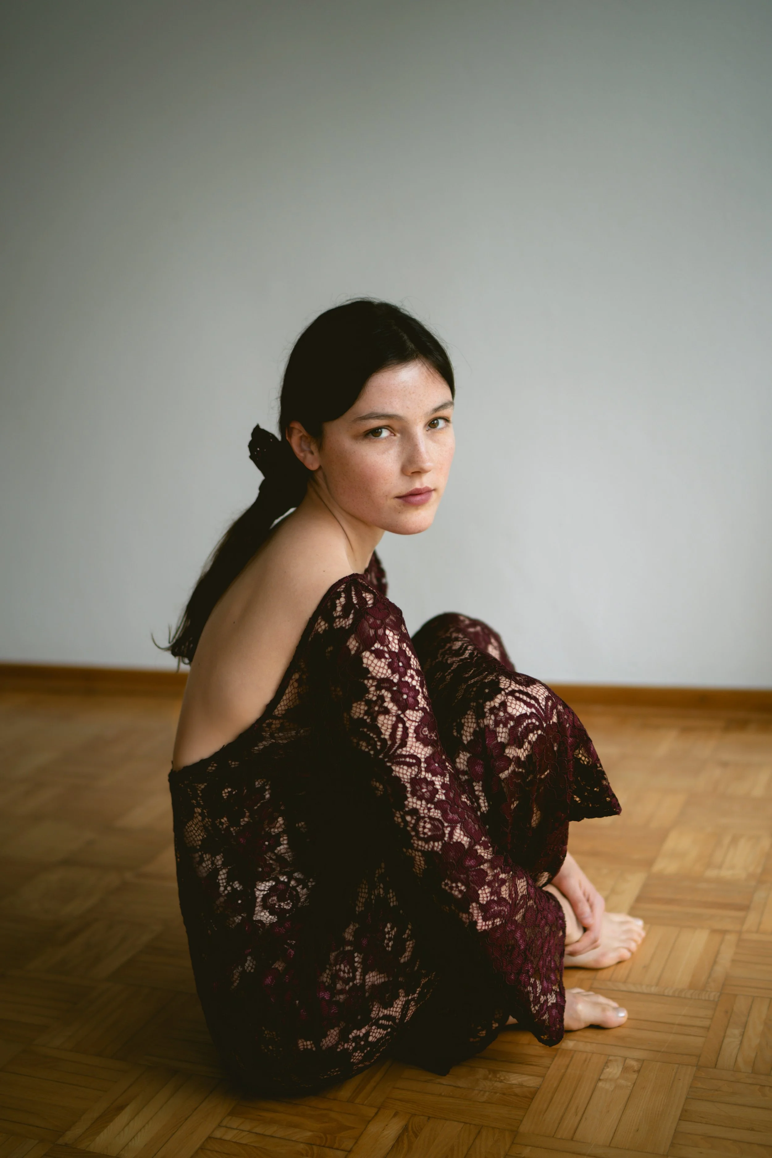 Ein junges Mädchen sitzt auf einem Holzboden, trägt ein rotes Spitzenkleid und schaut direkt in die Kamera.