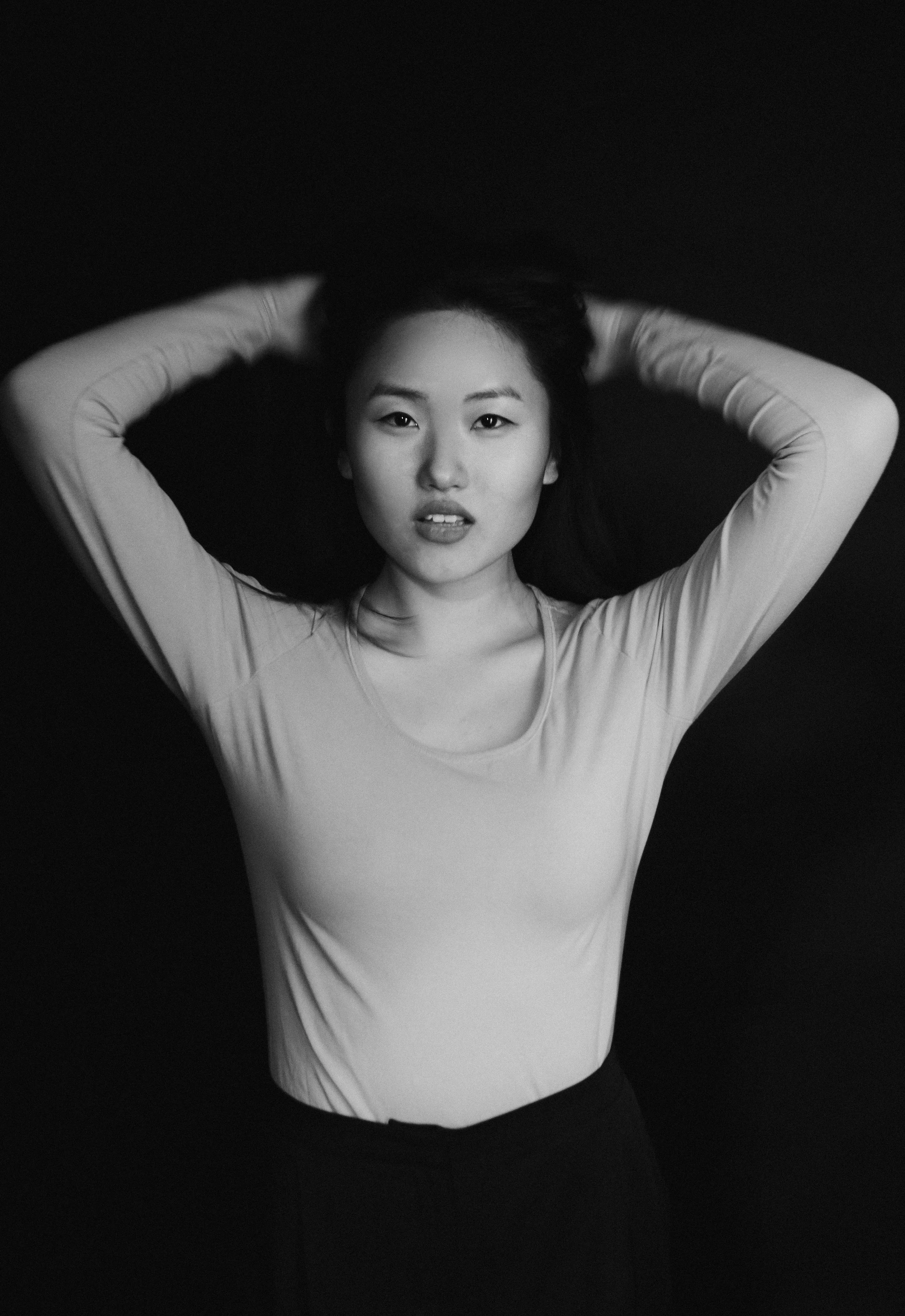 Schwarz-weiß-Fotografie eines jungen asiatischen Mädchens, das mit erhobenen Armen den Kopf hinter den Händen hält, vor einem dunklen Hintergrund.