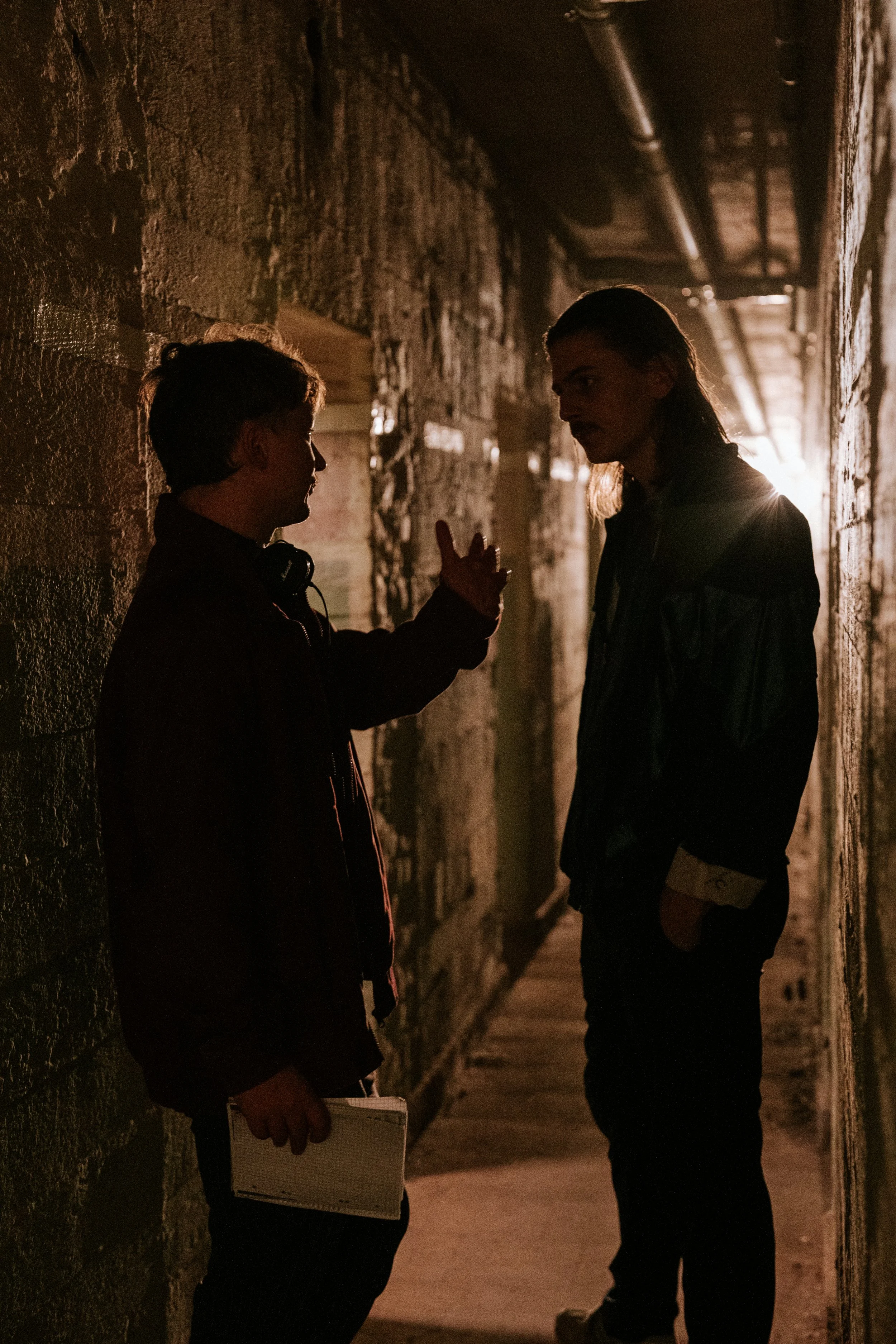 Zwei Menschen in einem dunklen, steinernen Korridor im Gespräch, eine Person zeigt mit der Hand nach oben, die andere hält ein Notizbuch.