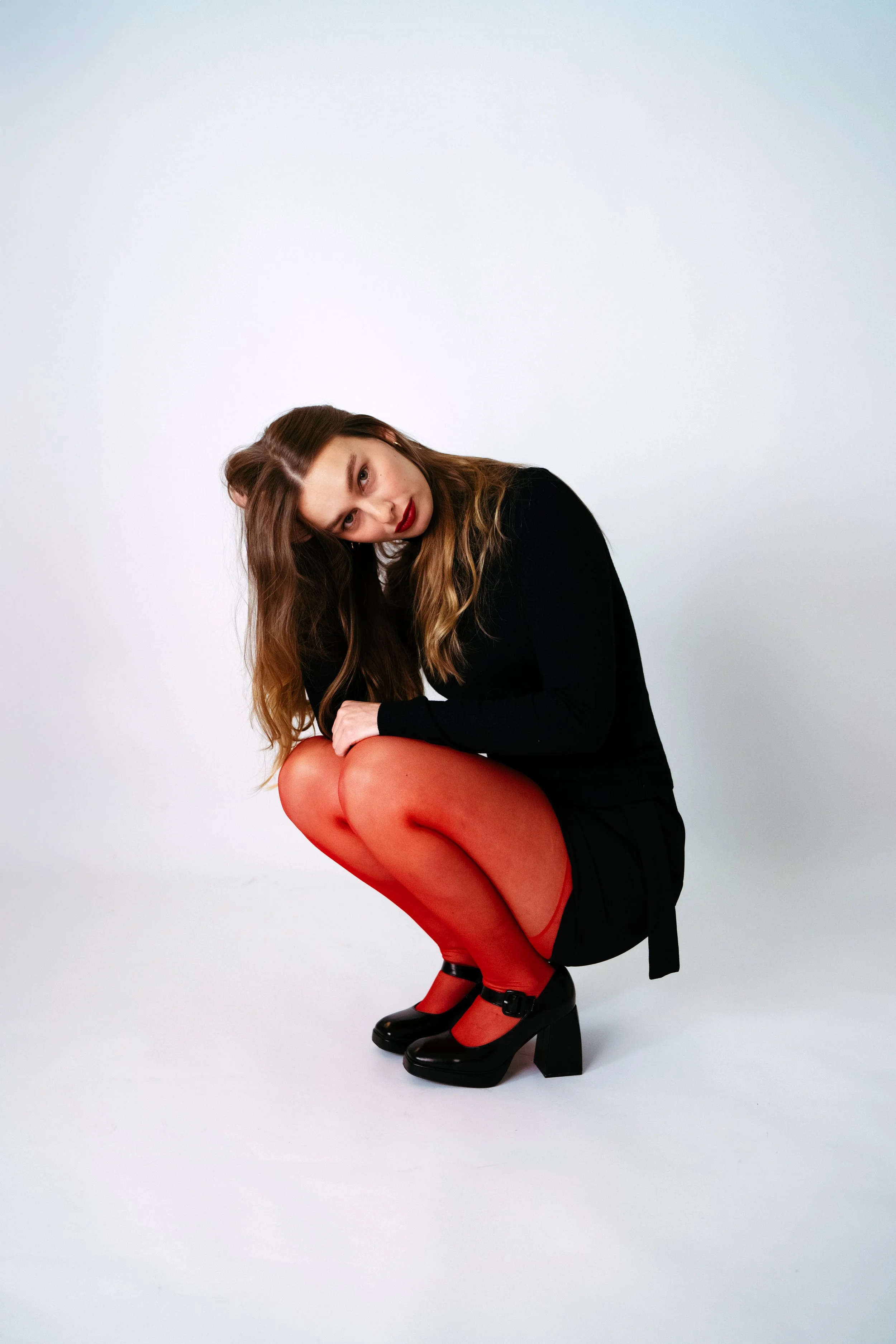 Junge Frau in Schwarz mit roten Pantoffeln, die in die Hocke geht und in die Kamera schaut.