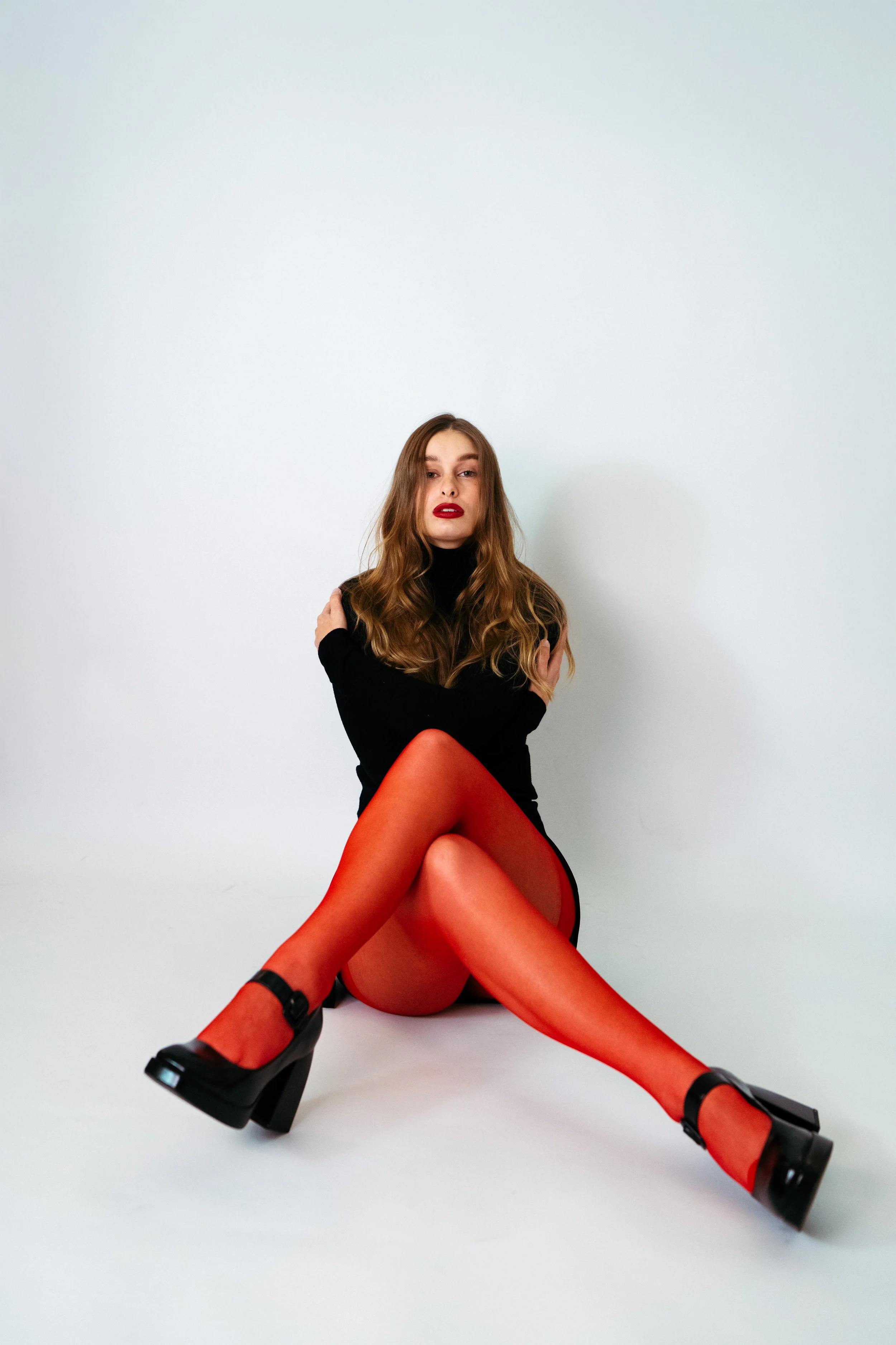 Ein Mädchen sitzt auf dem Boden vor einem weißen Hintergrund, trägt einen schwarzen Pullover, rote Strümpfe und schwarze Plateauschuhe, mit langem, gewelltem braunem Haar und rotem Lippenstift.