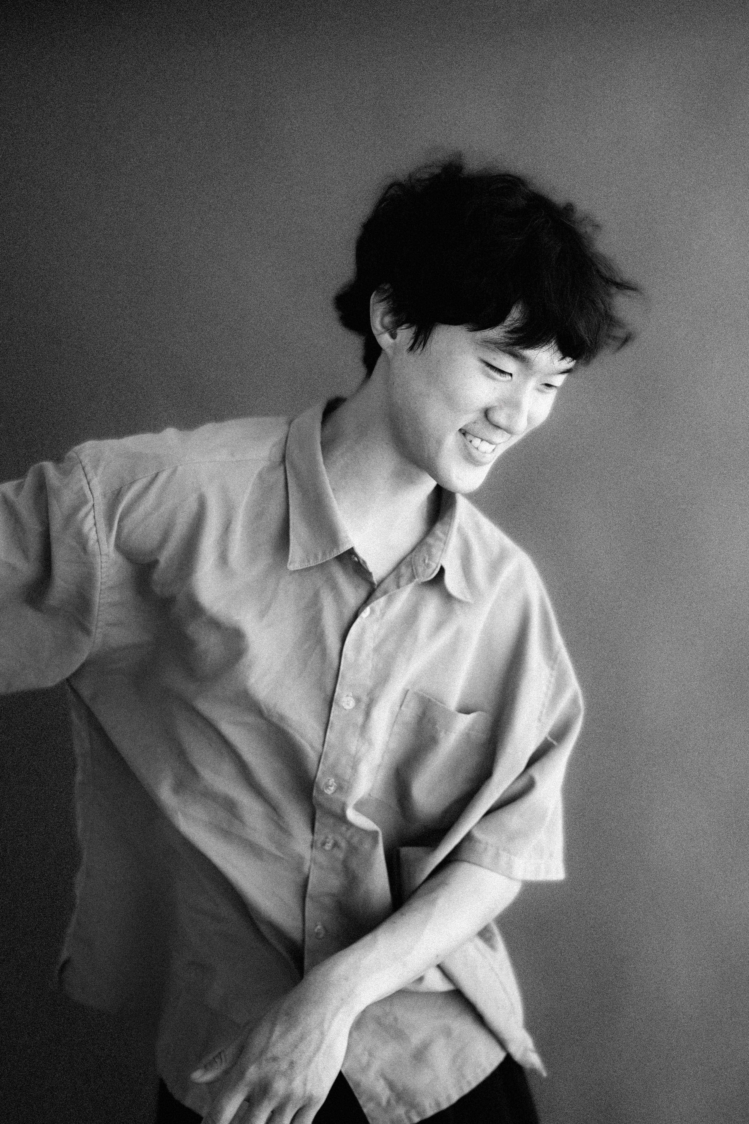 Junge Person mit dunklen, lockigen Haaren, lachend, in einem hellen Hemd, Schwarz-Weiß-Fotografie, lächelnd, von der Seite gesehen.