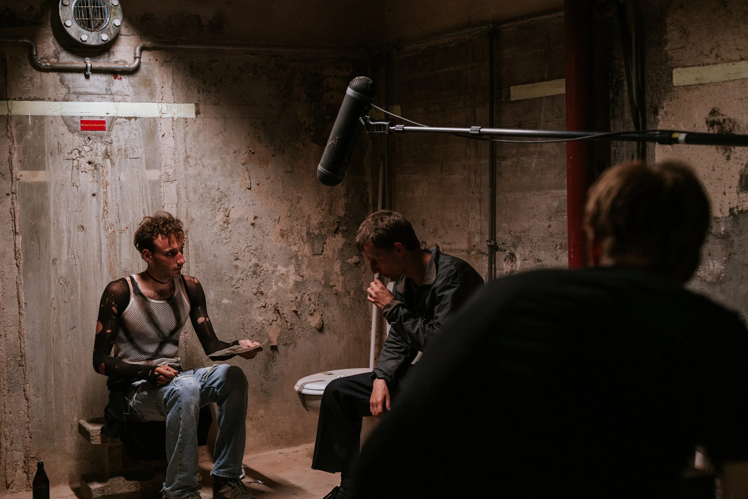 Drei junge Männer sitzen in einem rauen, unvollendeten Raum. Einer trägt ein durchlöchertes Oberteil, die anderen tragen dunkle Jacken. Ein Mikrofon hängt an einer langen Stange über ihnen, vermutlich für ein Interview oder eine Aufnahme.