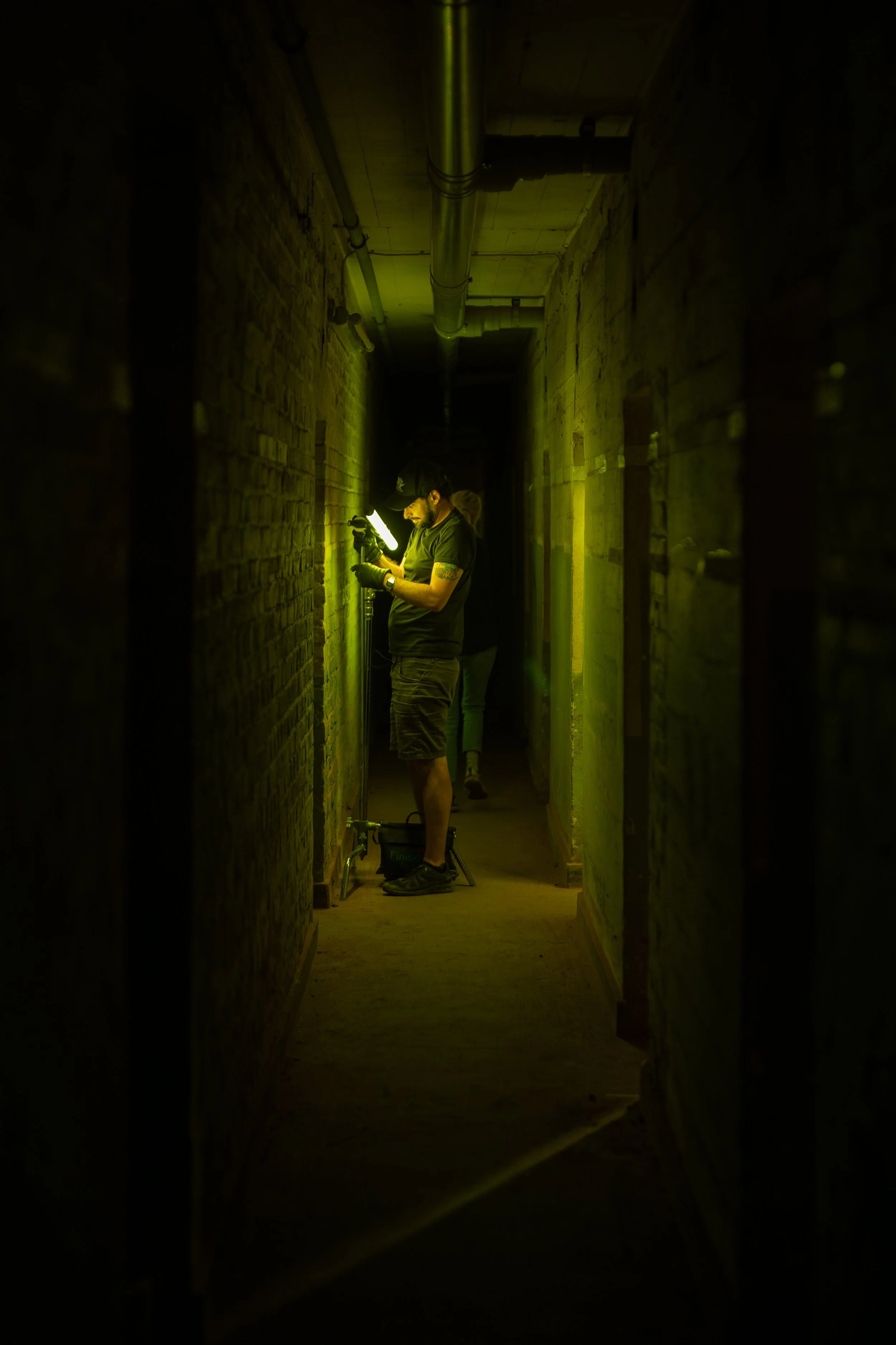 Mann in einem dunklen Flur hält eine Taschenlampe und an einem Rohr, trägt schwarze Kleidung, Basecap, Handschuhe und Shorts.