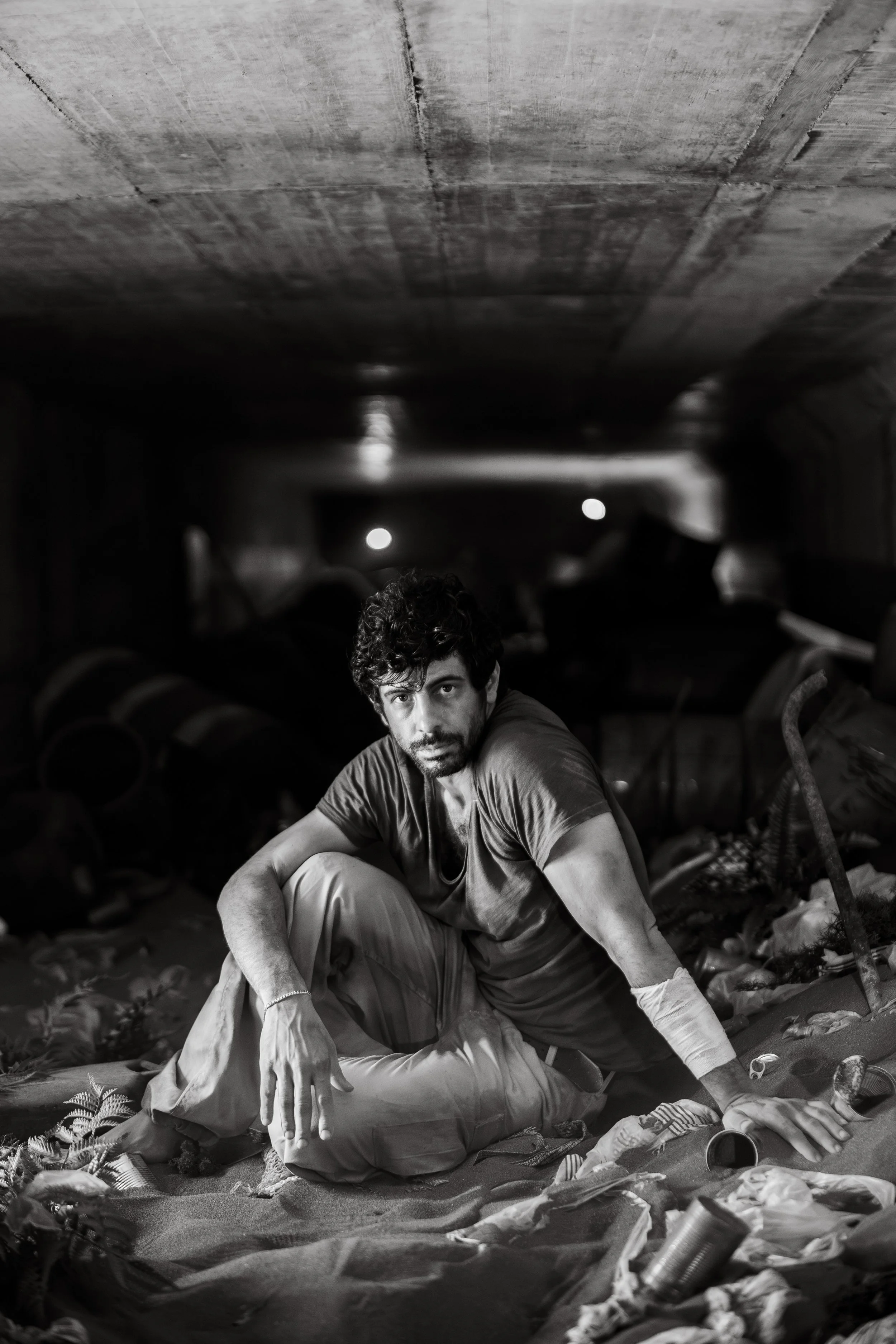 Schwarz-Weiß-Foto eines Mannes, der in einem dunklen, unordentlichen Raum auf dem Boden sitzt, umgeben von Müll und Abfällen.