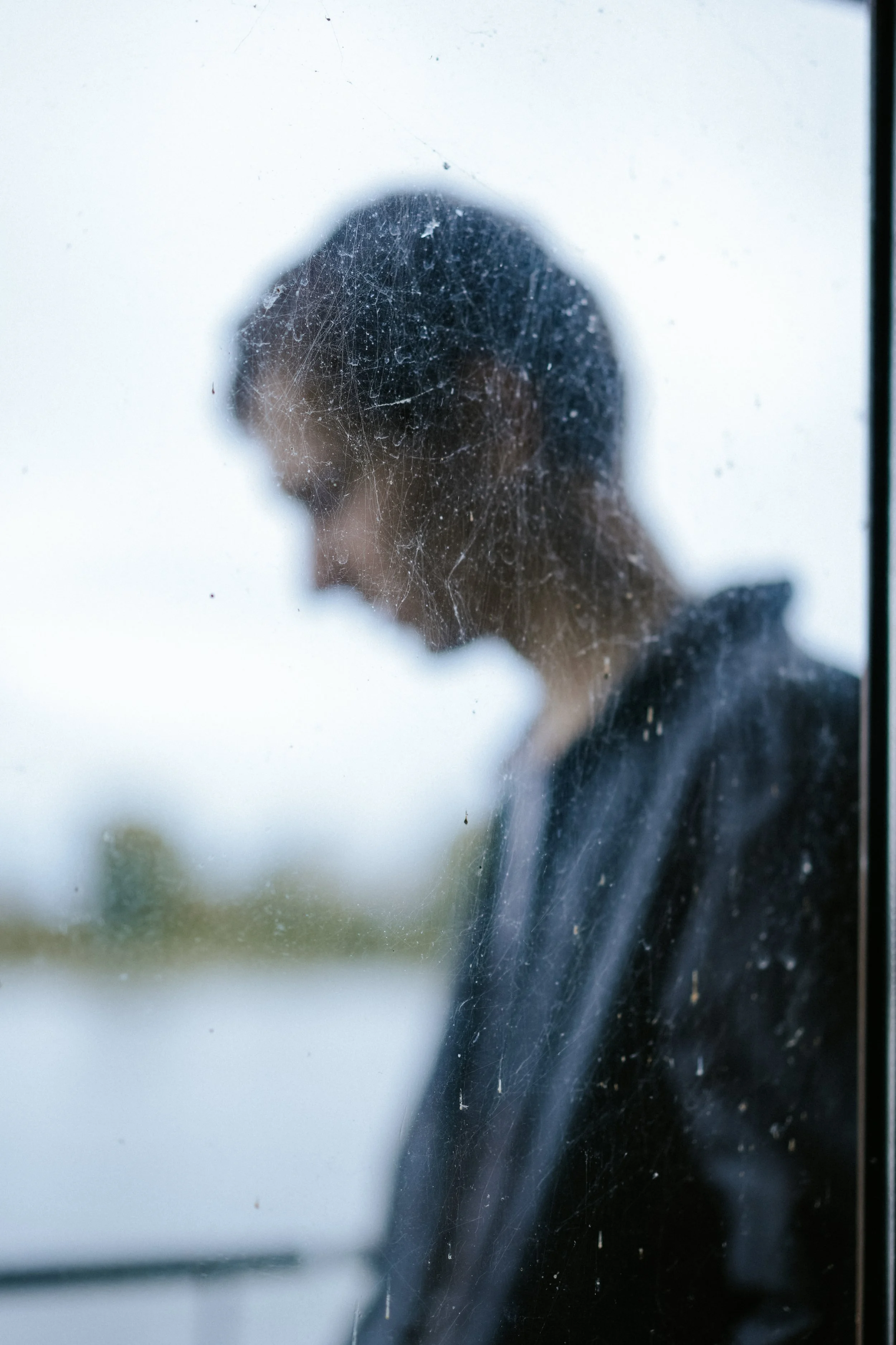 Der Blick durch ein schmutziges Fenster auf eine nachdenkliche Person, die nach links schaut. Im Hintergrund ist Wasser und eine verschwommene Natur zu sehen.