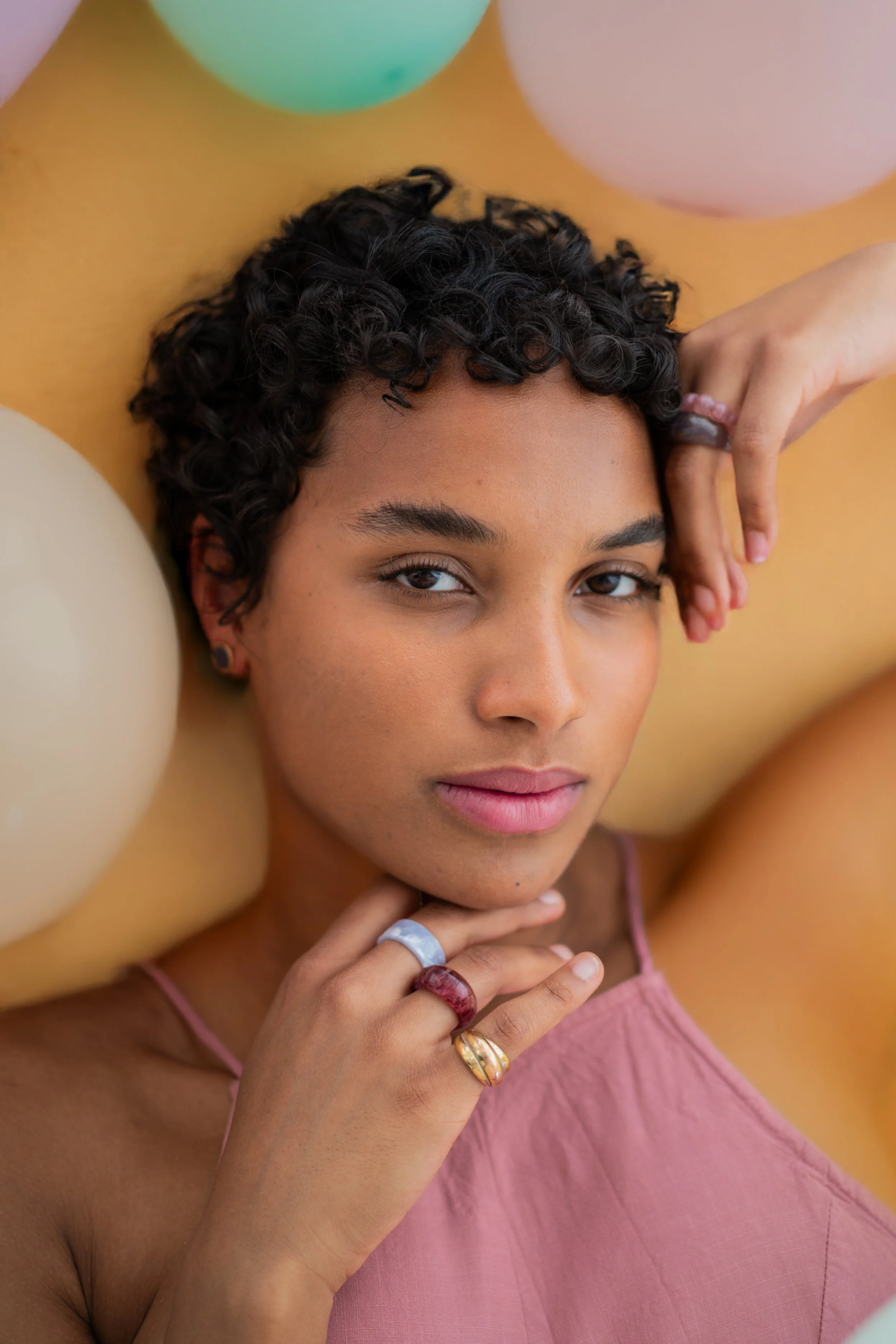 Junge Frau mit dunklen, lockigen Haaren und mehreren bunten Ringen, liegend auf gelbem Hintergrund, umgeben von pastellfarbigen Ballons.