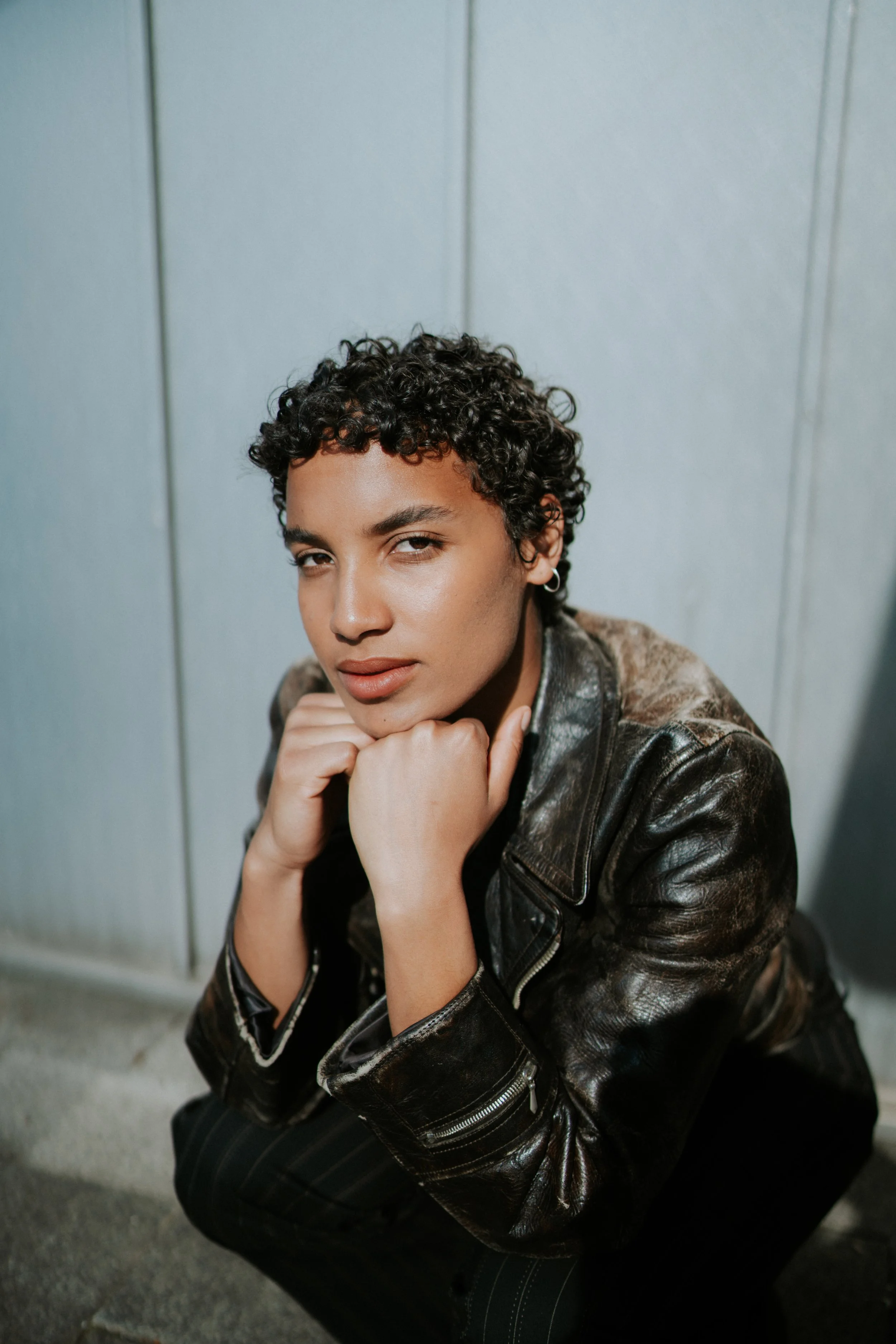 Junge Frau mit dunklen, lockigen Haaren, die ein dunkles Lederjacke trägt, sitzt auf dem Boden vor einer hellgrauen Wand und blickt nach oben, mit einer Hand unter dem Kinn.