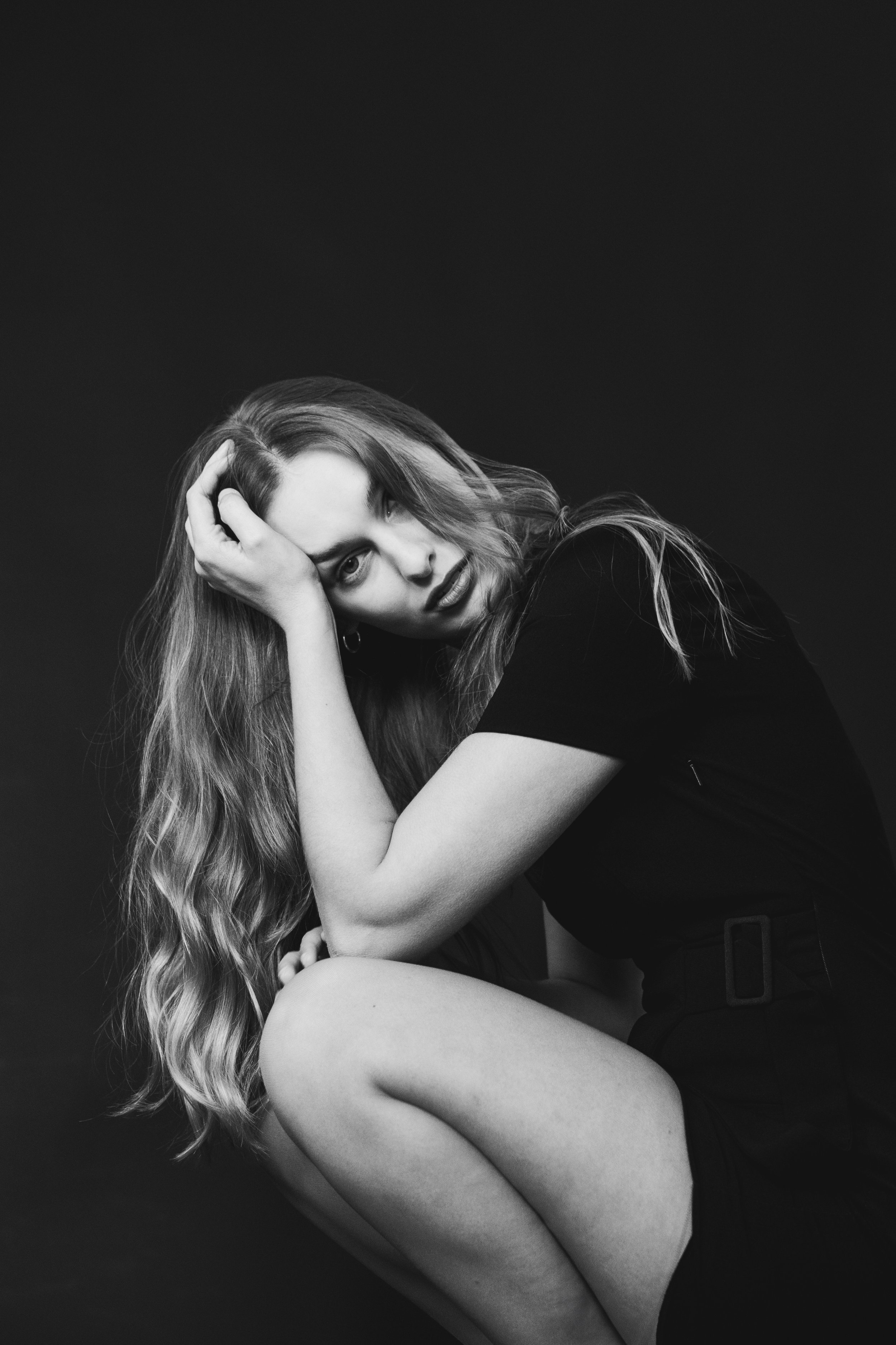 Schwarz-weiß-Fotografie eines nachdenklichen Mädchens mit langen, gewellten Haaren, sitzend mit Knie angezogen, in dunklem T-Shirt gegen dunklem Hintergrund.