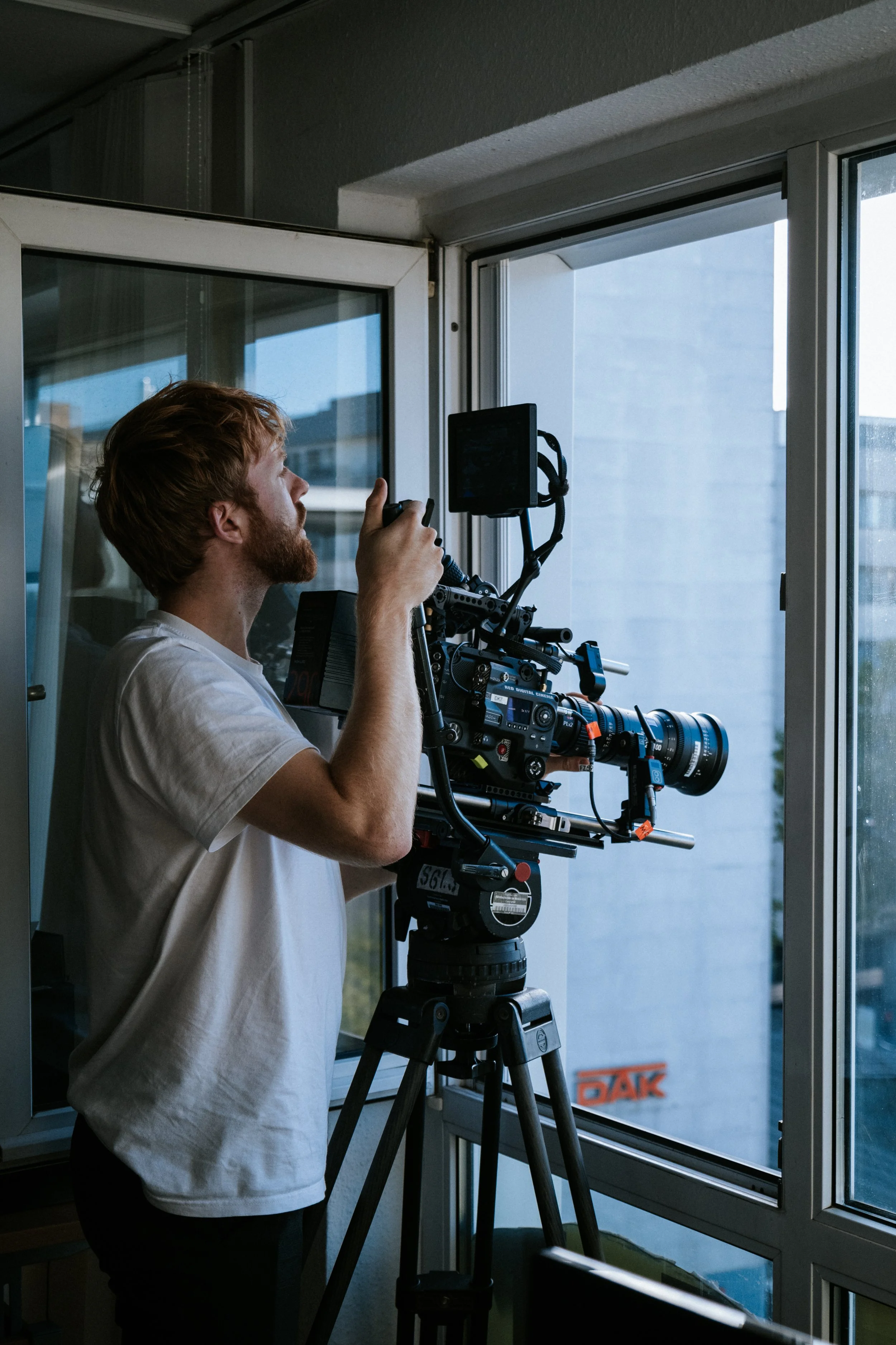 Ein Mann steht vor einem großen Fenster und bedient eine professionelle Filmkamera auf einem Stativ, während er in die Kamera hineinschaut.