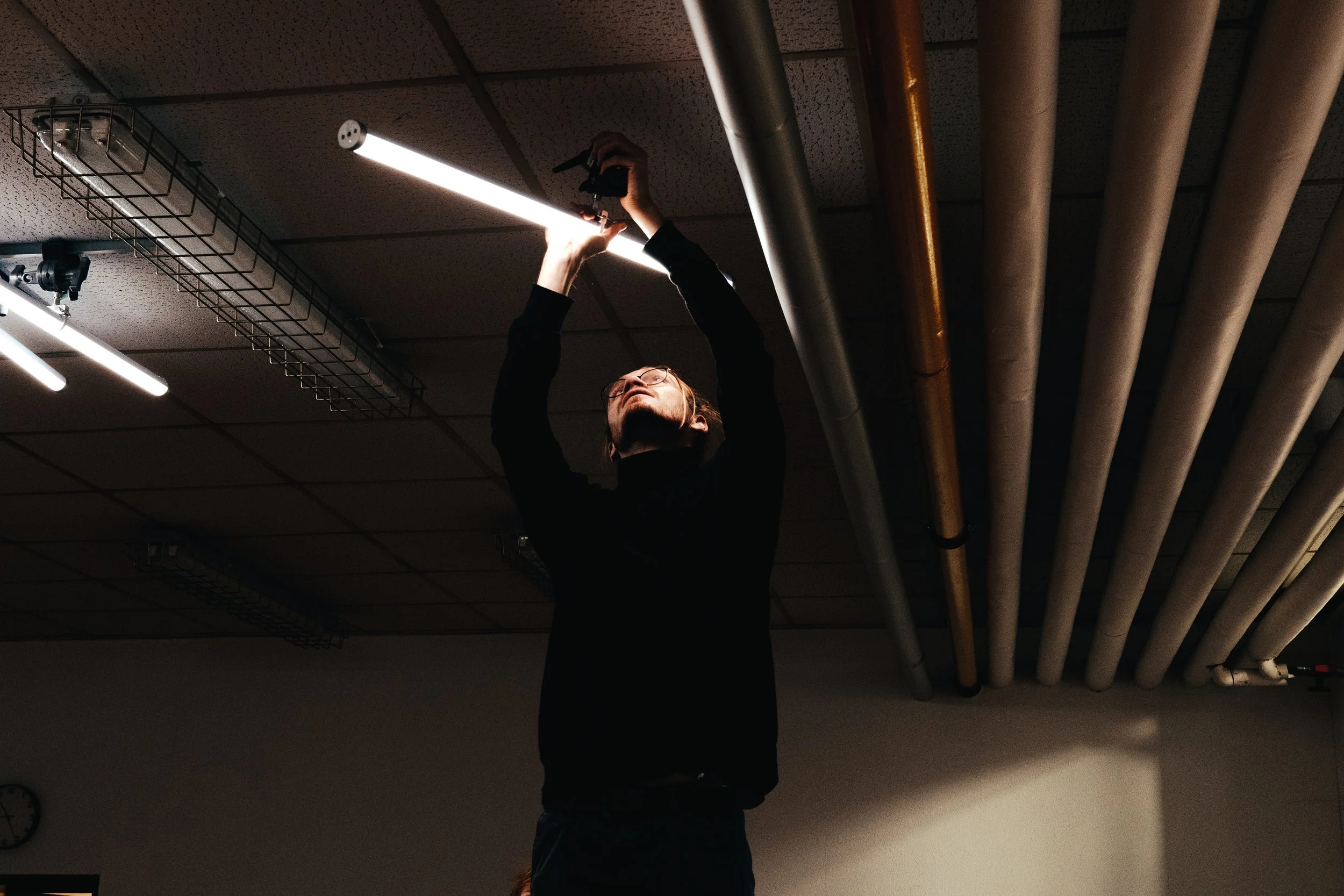 Person in dunklem Oberteil installiert eine Lichtleiste an der Decke in einem Raum mit sichtbaren Rohrleitungen.