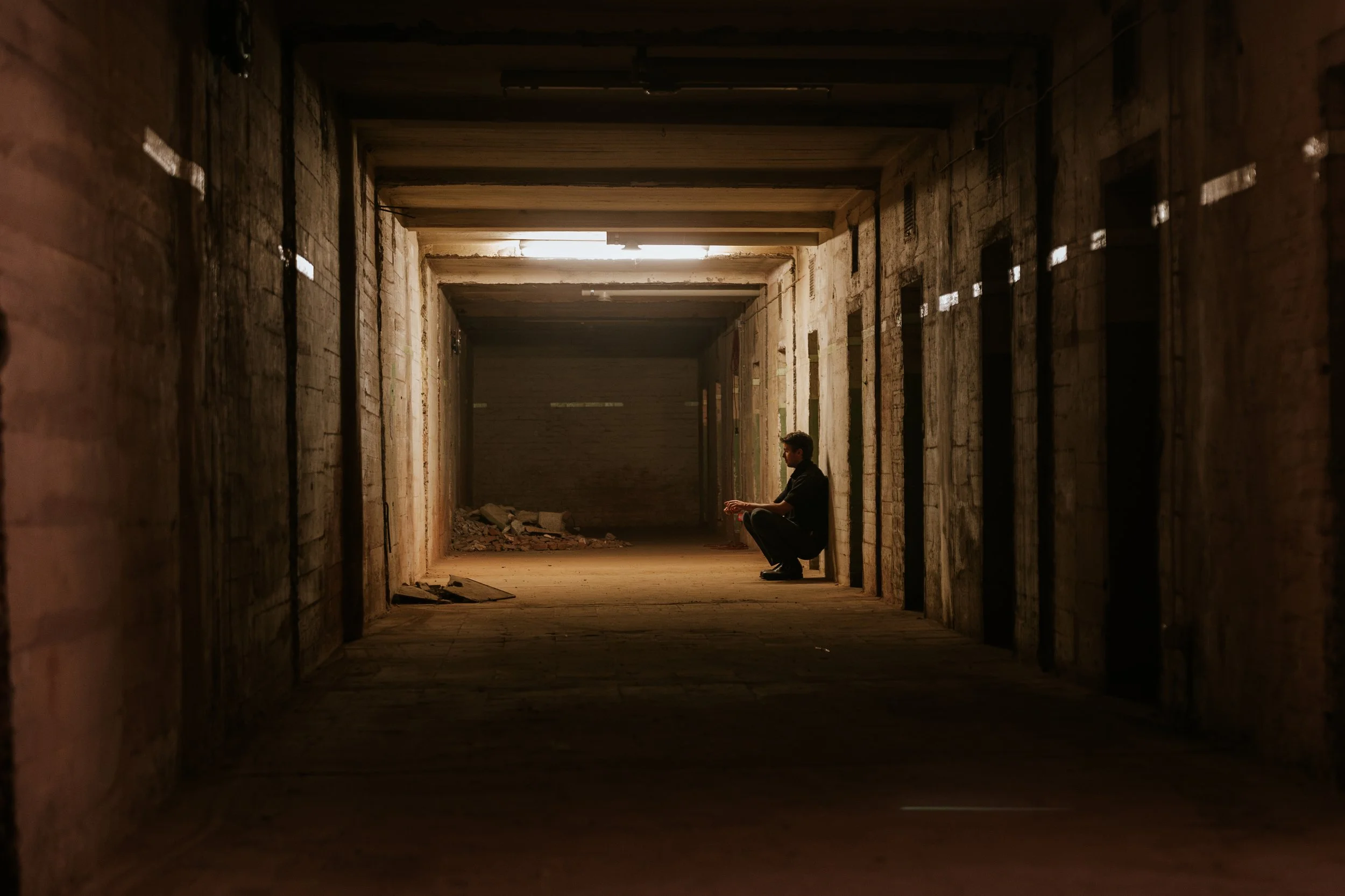 Ein junger Mann sitzt in einem dunklen, verlassenen Gebäude in einer Ecke, umgeben von steinernen Wänden und Betonsäulen, mit gedämpftem Licht, das von oben scheint.