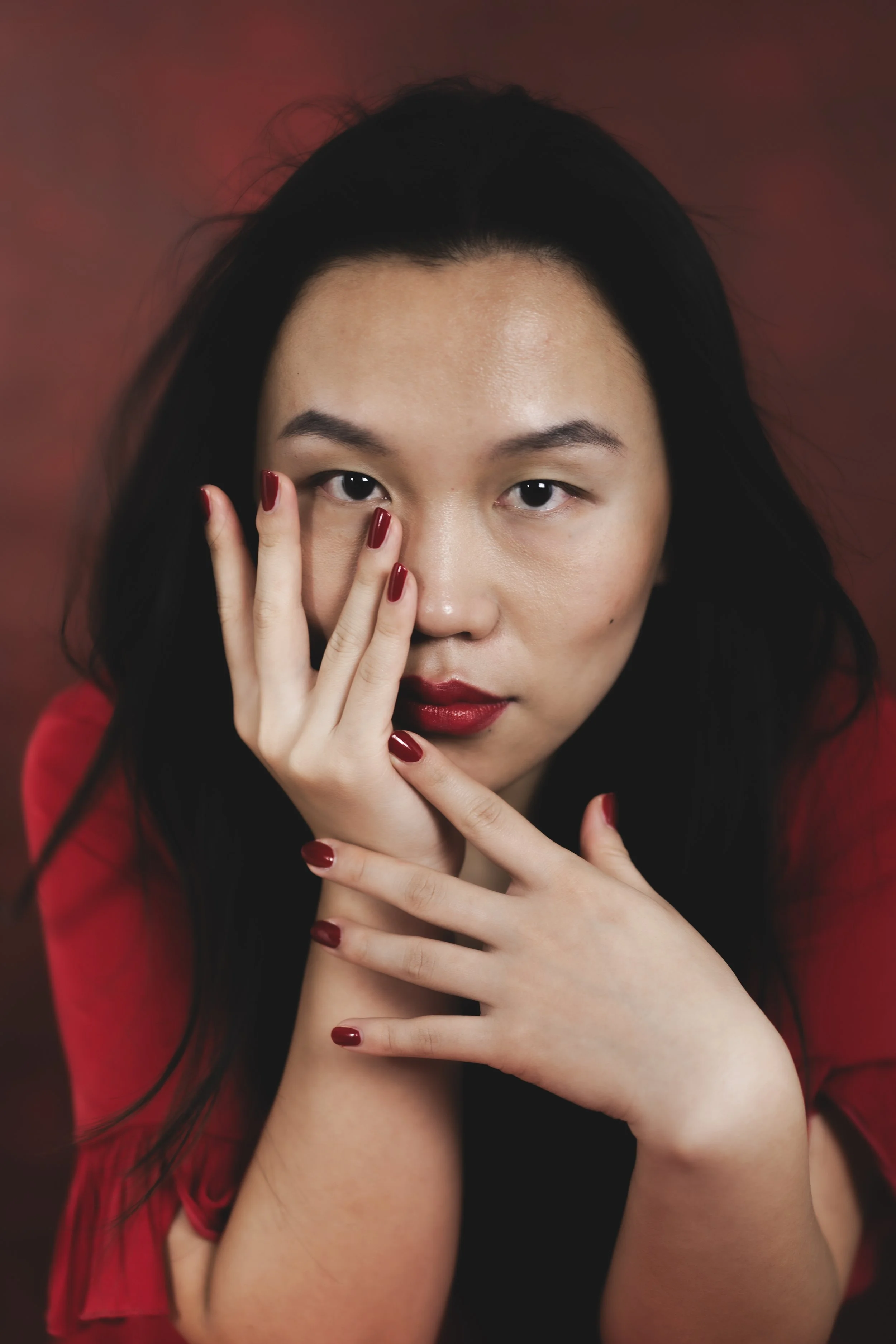 Porträt einer asiatischen Frau mit dunklem Haar, roten Lippen und roten Nägeln, die ihre Hand vor das Gesicht hält, vor einem dunklen, warmfarbenen Hintergrund.