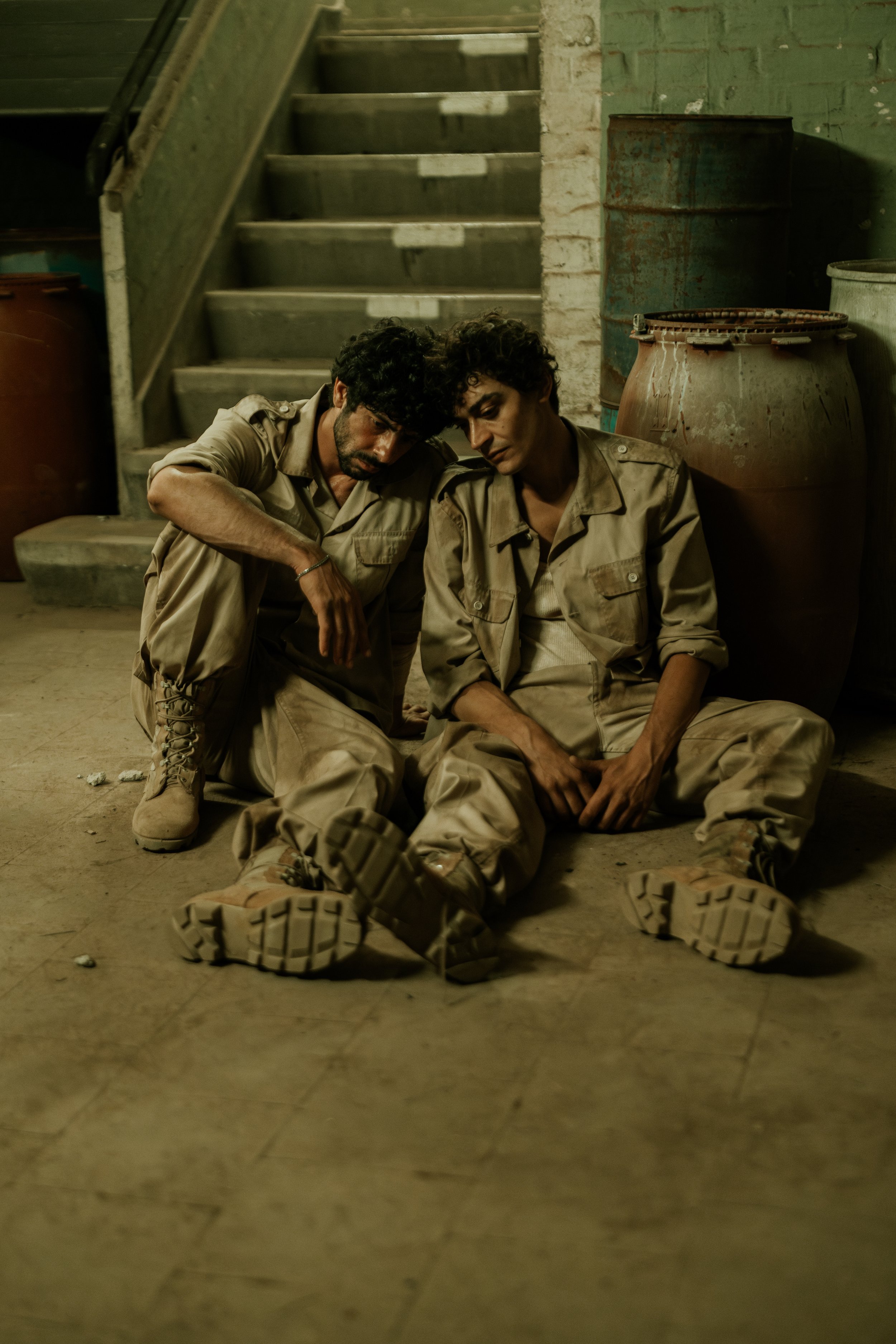 Zwei Männer in Khaki-Uniformen sitzen erschöpft am Boden vor einem grünen Wand, mit Treppe im Hintergrund und großen Tonkrügen neben ihnen.