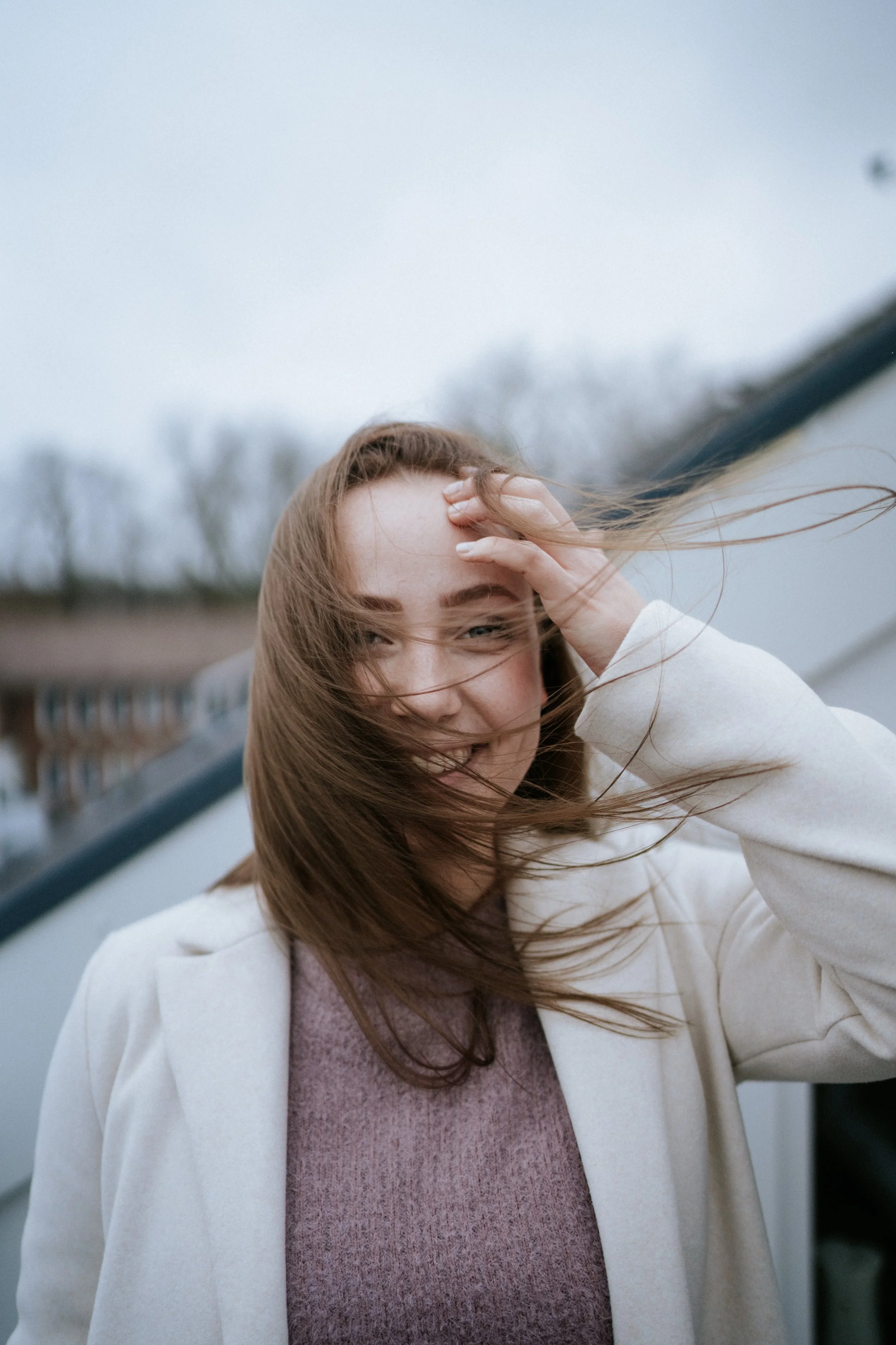 Junge Frau mit langen braunen Haaren, lacht im Wind, trägt einen weißen Mantel und einen lila Pullover, draußen bei bewölktem Himmel.