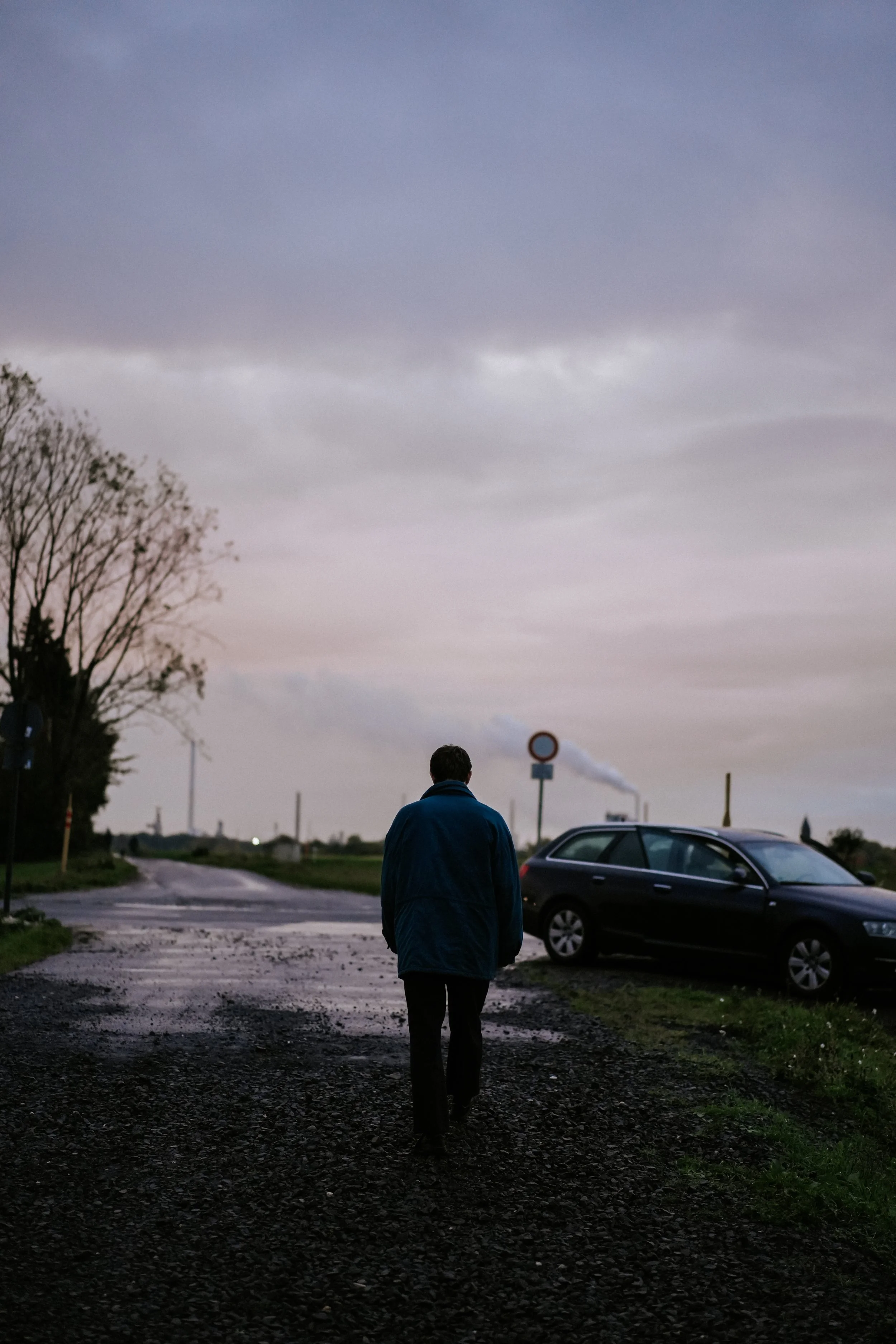 Ein Mann läuft alleine auf einem schmutzigen Weg bei bewölktem Himmel, neben einem Auto. Rauch steigt aus einer Fabrik in der Ferne.