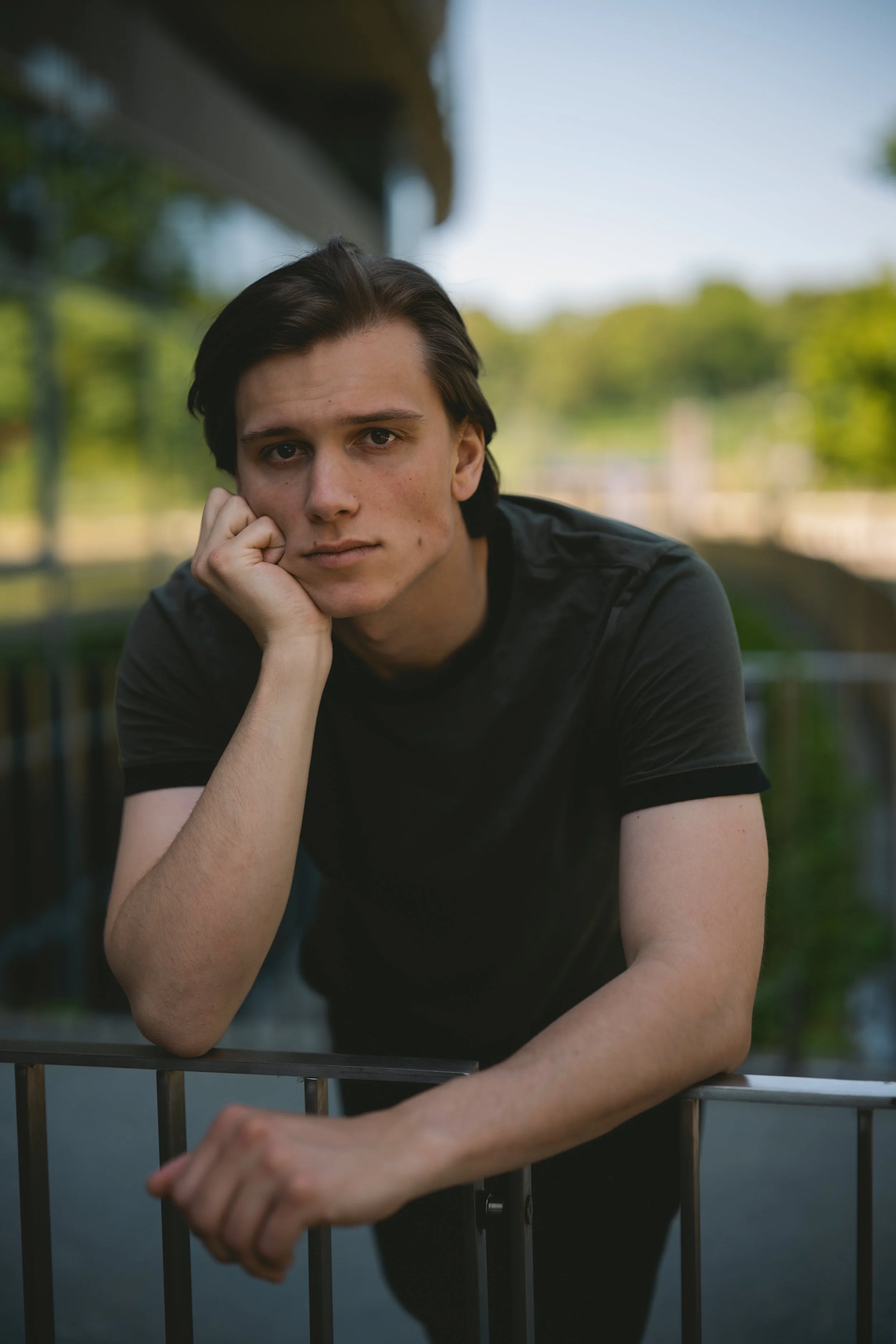 Junger Mann lehnt mit seinem Arm auf einem Metallzaun, schaut nachdenklich in die Kamera, umgeben von grüner Natur bei Tageslicht.