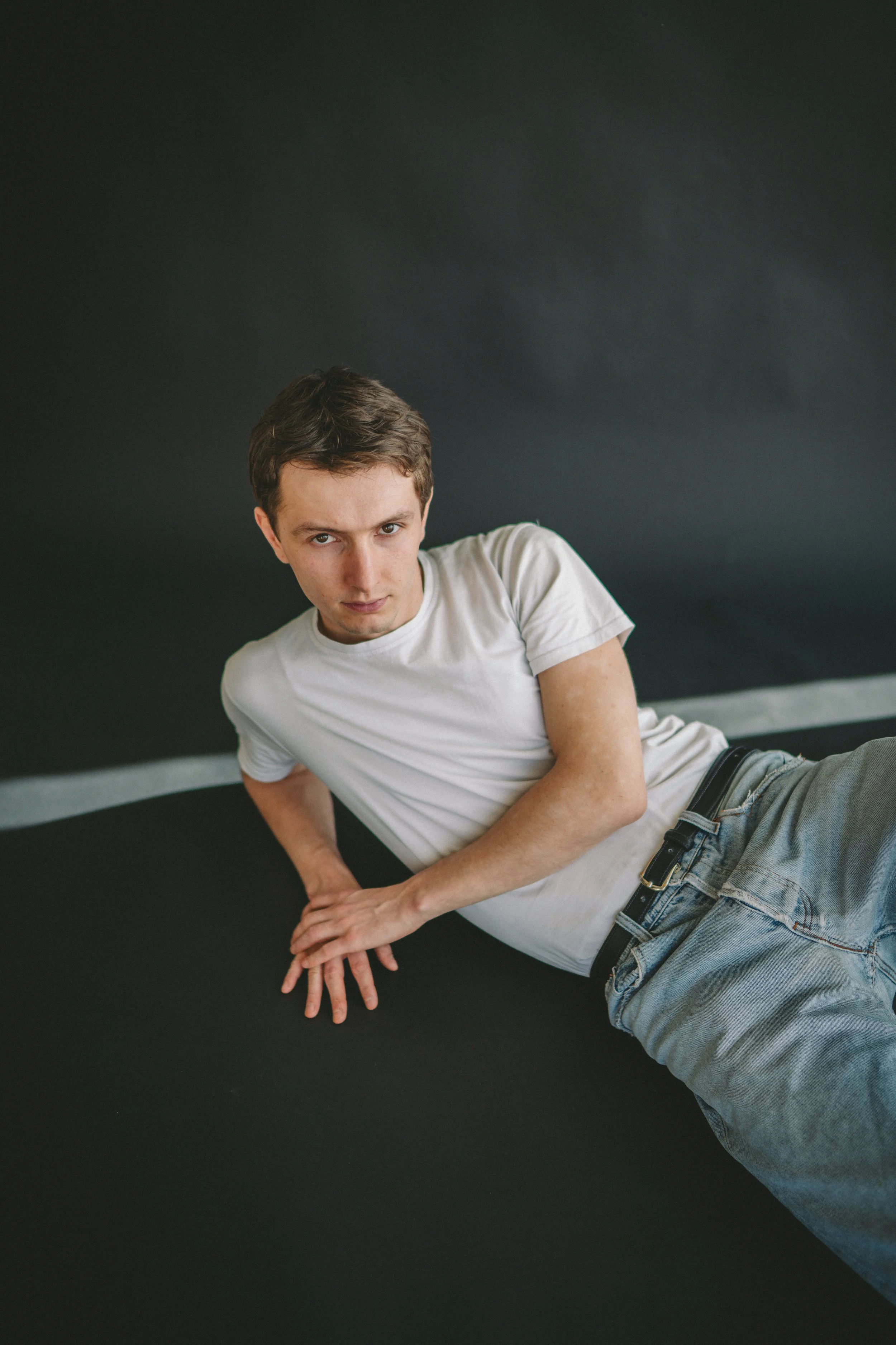 Junger Mann liegt seitlich auf einem schwarzen Boden, schaut direkt in die Kamera, trägt weißes T-Shirt und Jeans.