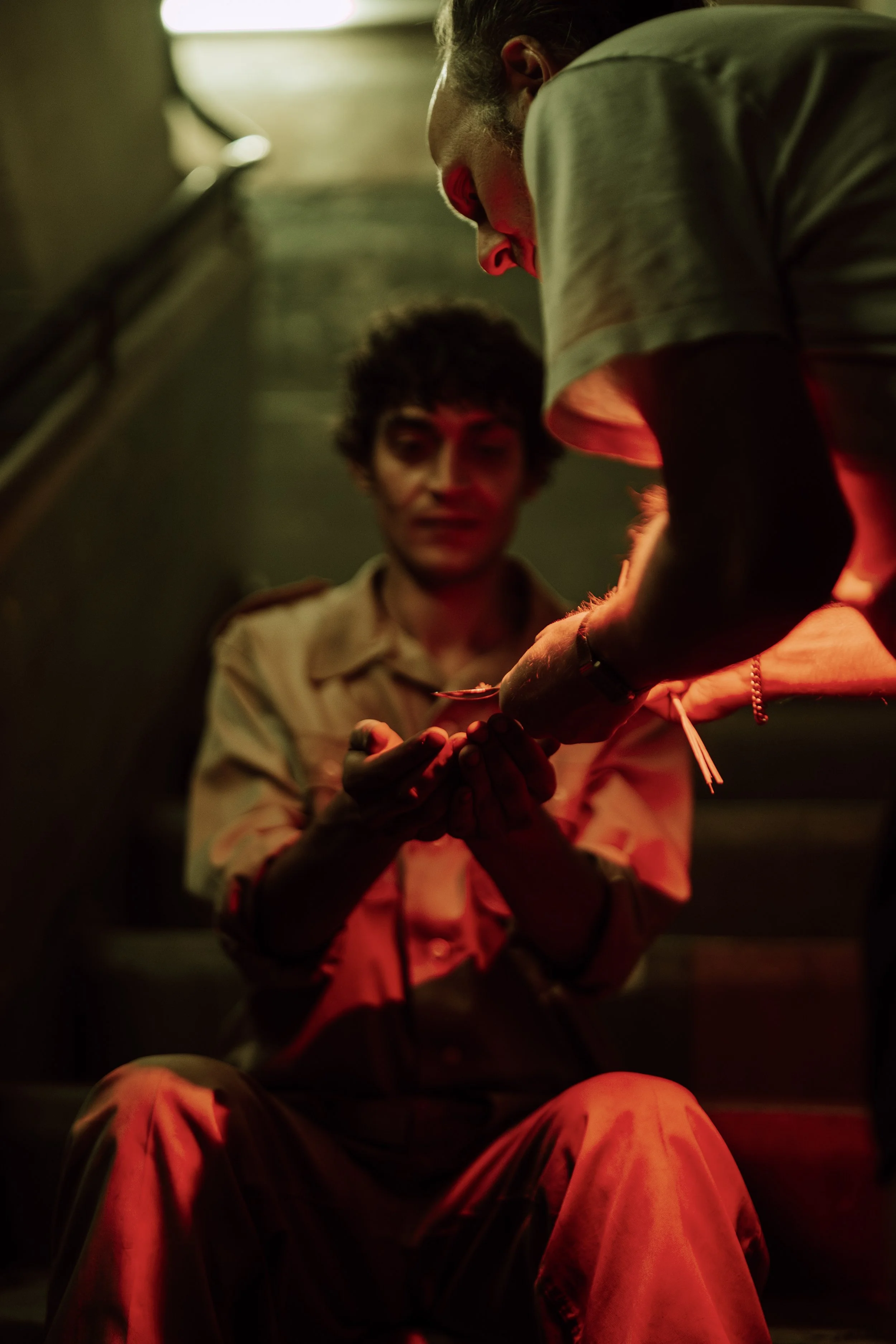 Zwei Menschen auf einer Treppe, einer sitzt und der andere steht, bei Nacht, im Schatten, mit rotem Licht. Der stehende Mensch reicht dem sitzenden Mensch etwas.