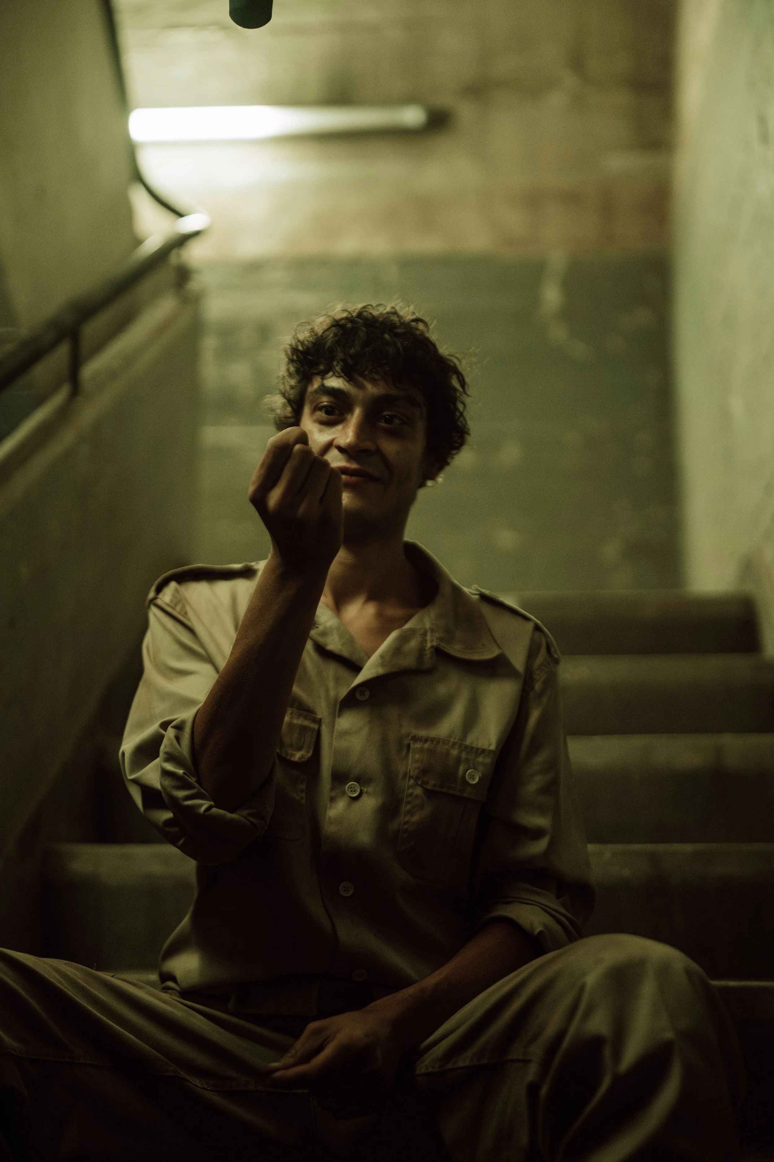 Ein junger Mann mit lockigem Haar sitzt auf einer Treppe in einem Gebäude mit grellem Licht im Hintergrund.
