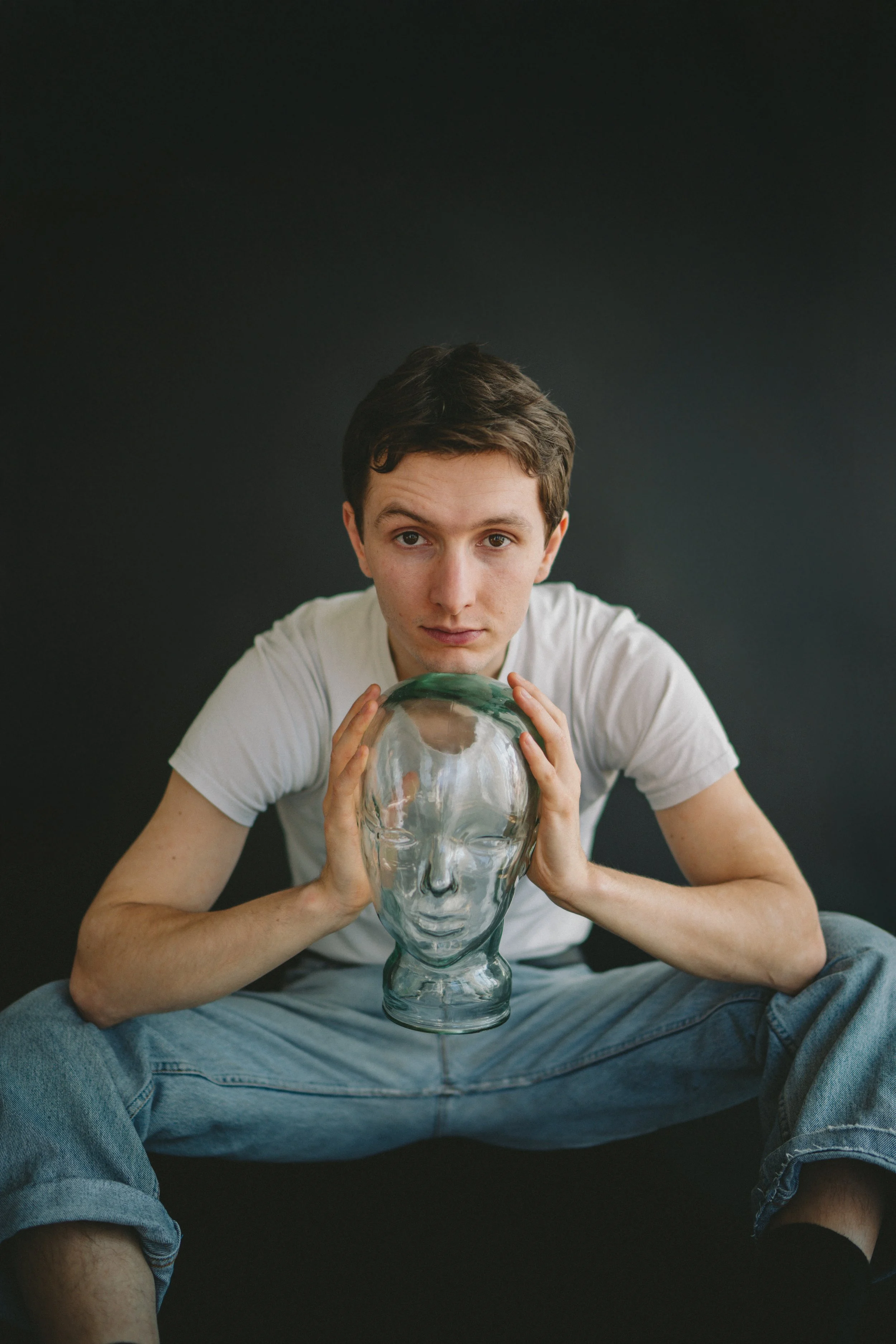 Junge Person sitzt auf dem Boden vor schwarzem Hintergrund und hält eine durchsichtige Glasvase in Form eines Kopfes. Er trägt ein weißes T-Shirt und Jeans und blickt direkt in die Kamera.