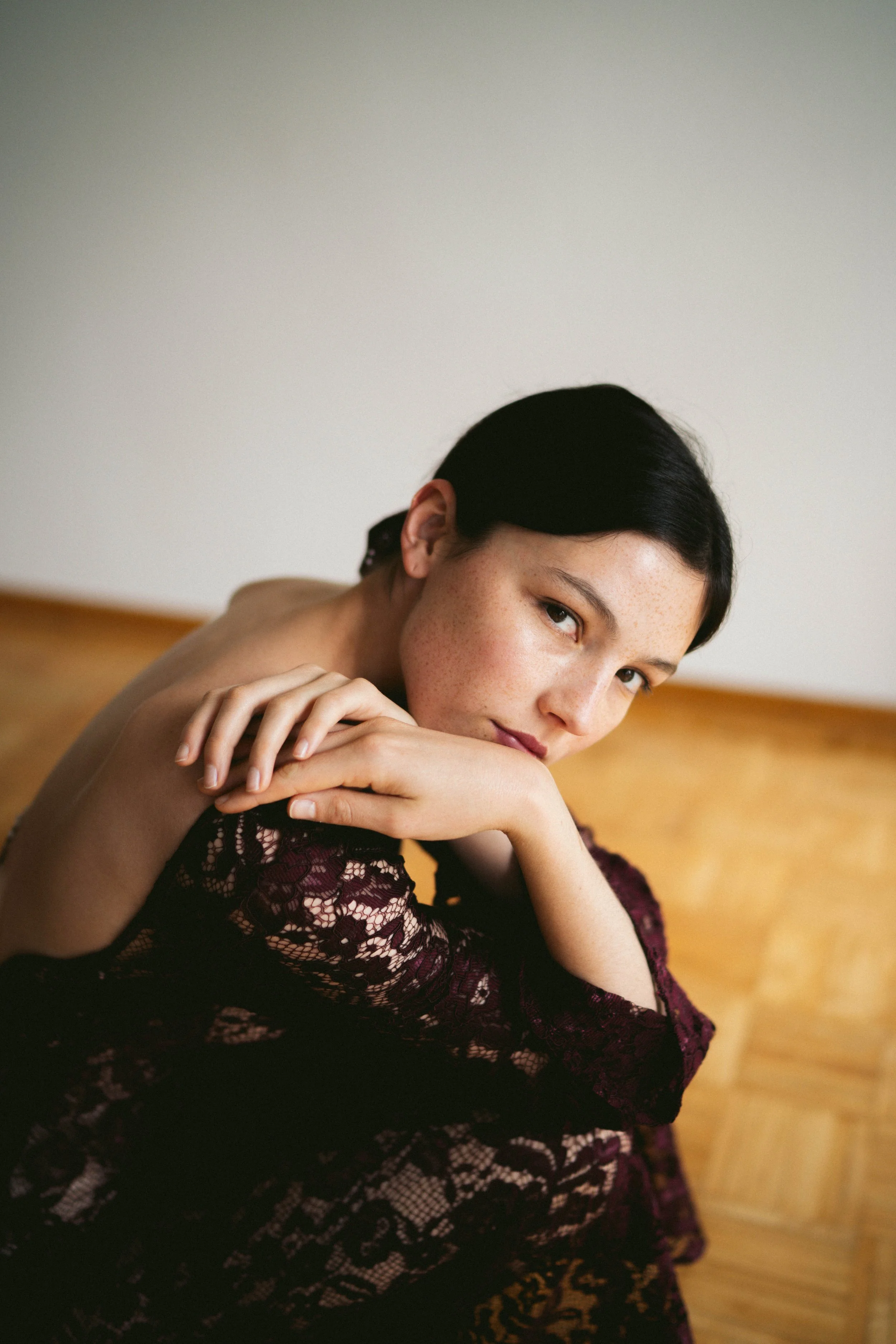 Ein junges Mädchen mit schwarzen Haaren und Sommersprossen, das nachdenklich in die Kamera schaut, während sie eine dunkle, violette Spitzenjacke trägt, auf einem Holzboden sitzend.