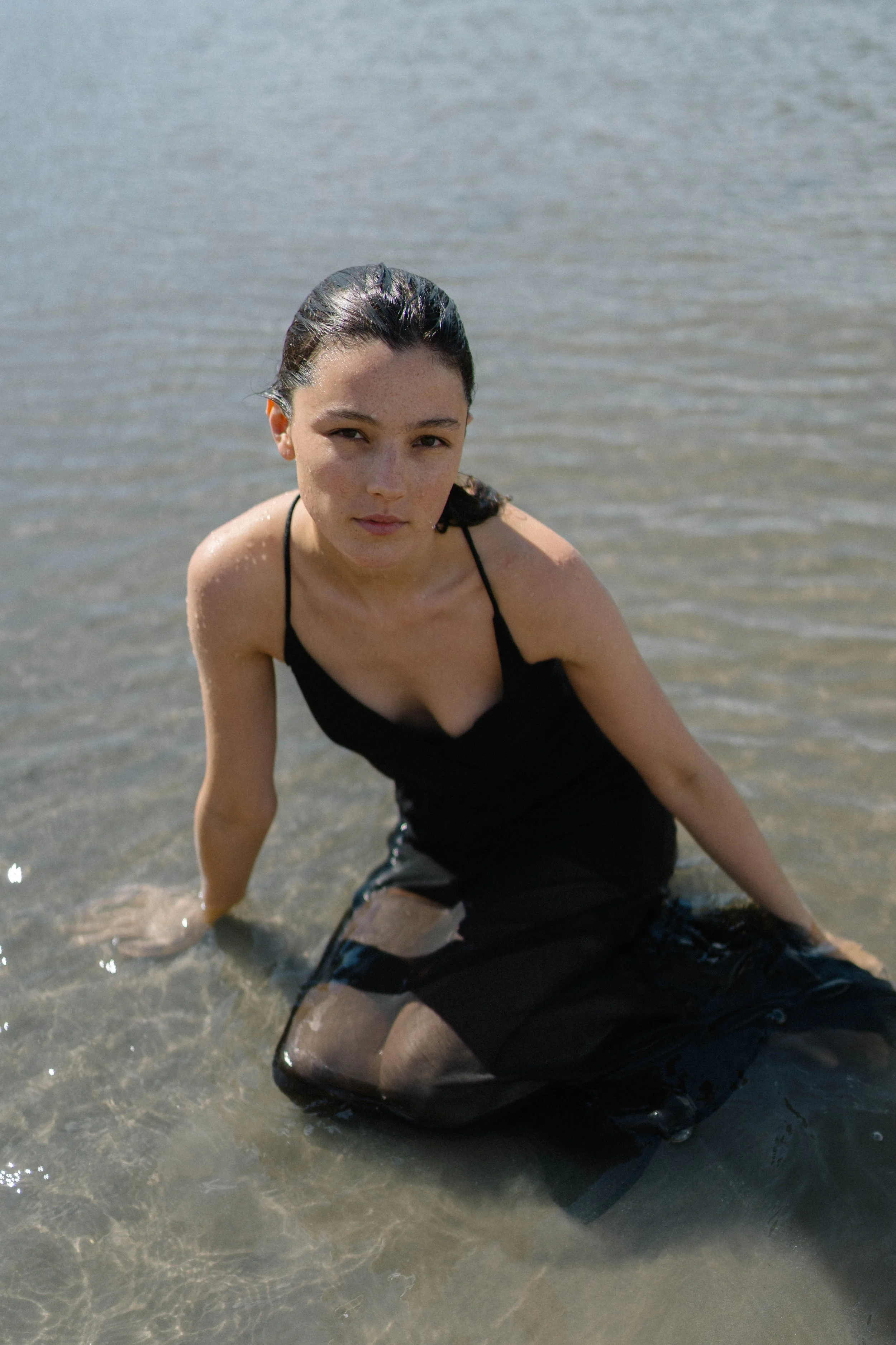 Junge Frau in schwarzem Rock sitzt im Wasser am Strand, Blick in die Kamera.