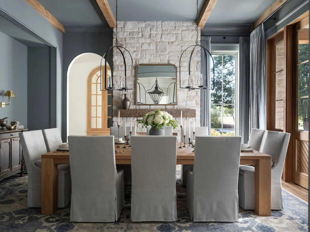 Dining room with wooden table, beige chairs, stone fireplace, large mirror, and large windows with curtains.