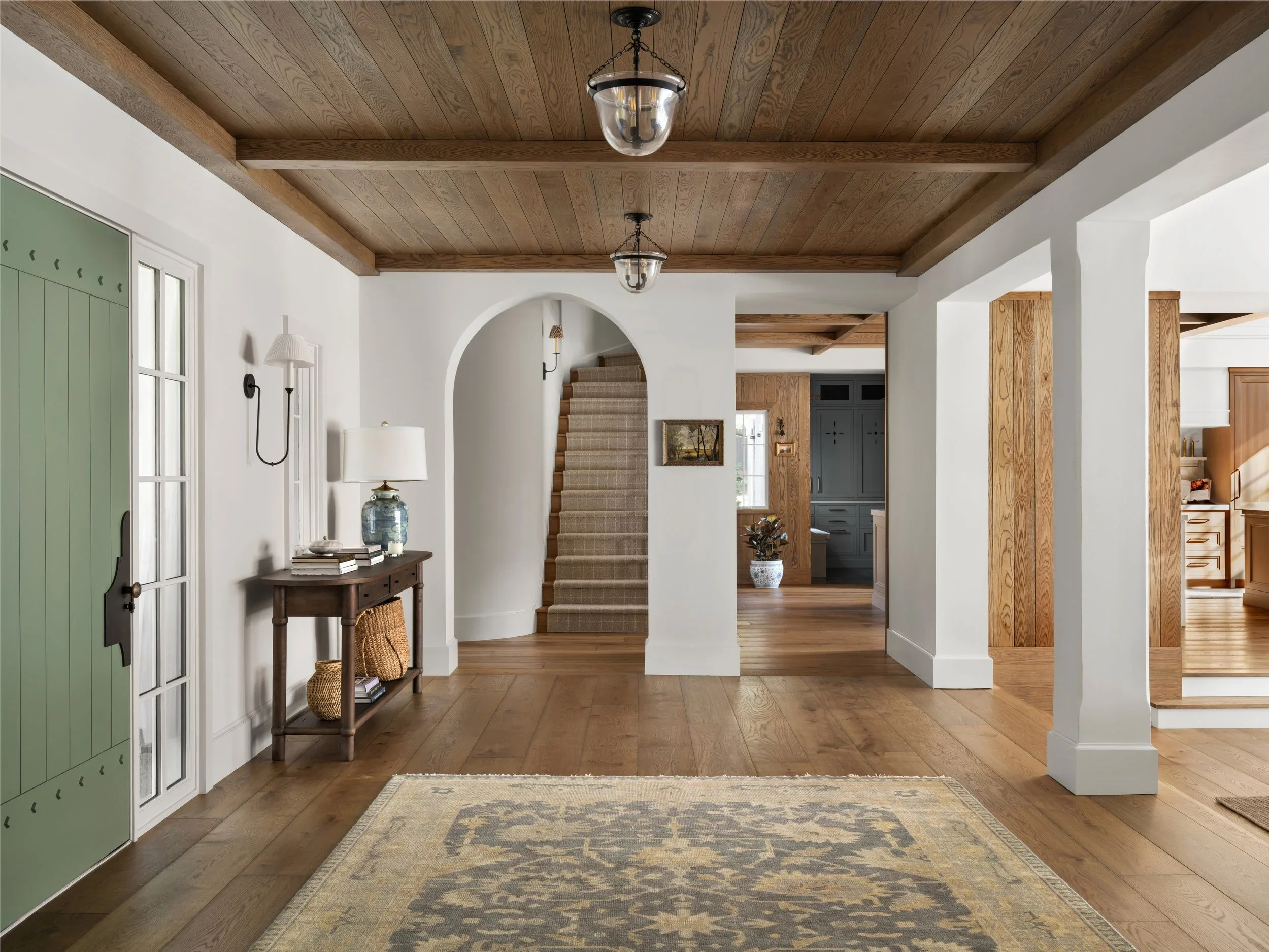 Entryway with wooden ceiling beams, dark hanging light fixtures, light-colored walls, a small console table with decorative items, a patterned rug, and an arched doorway leading to a staircase and another room.
