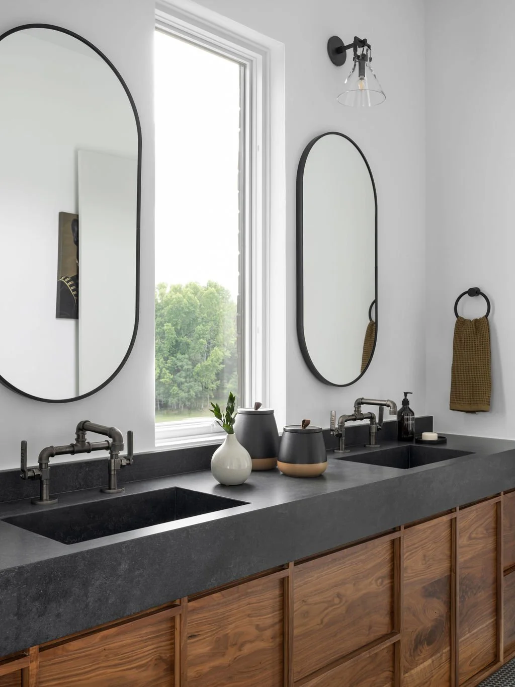 A modern bathroom vanity with a dark countertop, double black sinks, two oval mirrors, a window, and decorative vases and accessories.