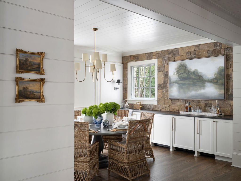 A dining room with a round table surrounded by wicker chairs, a chandelier hanging from the ceiling, a window on a stone accent wall, and a painting of a river scene on the wall.