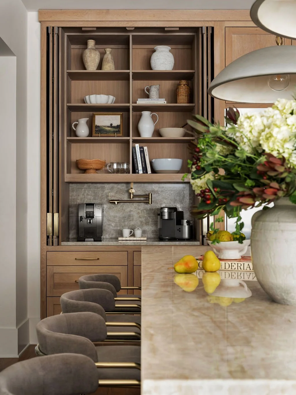 Modern kitchen with a light wood open shelving unit displaying decorative vases, bowls, and books, a coffee station with espresso machine, and a large flower arrangement with pears on a polished marble countertop with bar stools.