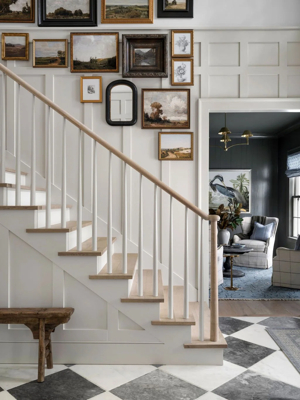 Living room with white staircase, gallery wall of landscape paintings, and seating area with armchairs and artwork of a heron.