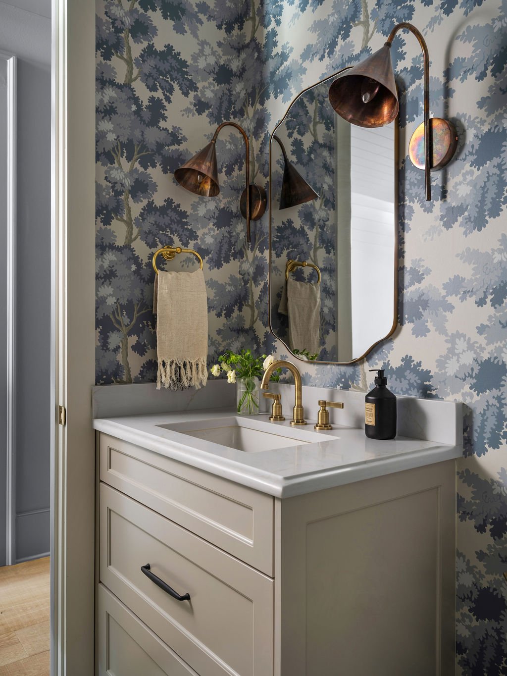 A bathroom vanity with a white countertop, gold faucet, and beige cabinet, decorated with a vase of white flowers. A large irregular mirror hangs on the wall above, which is covered in blue and gray leafy wallpaper. Copper cone-shaped wall sconces wi