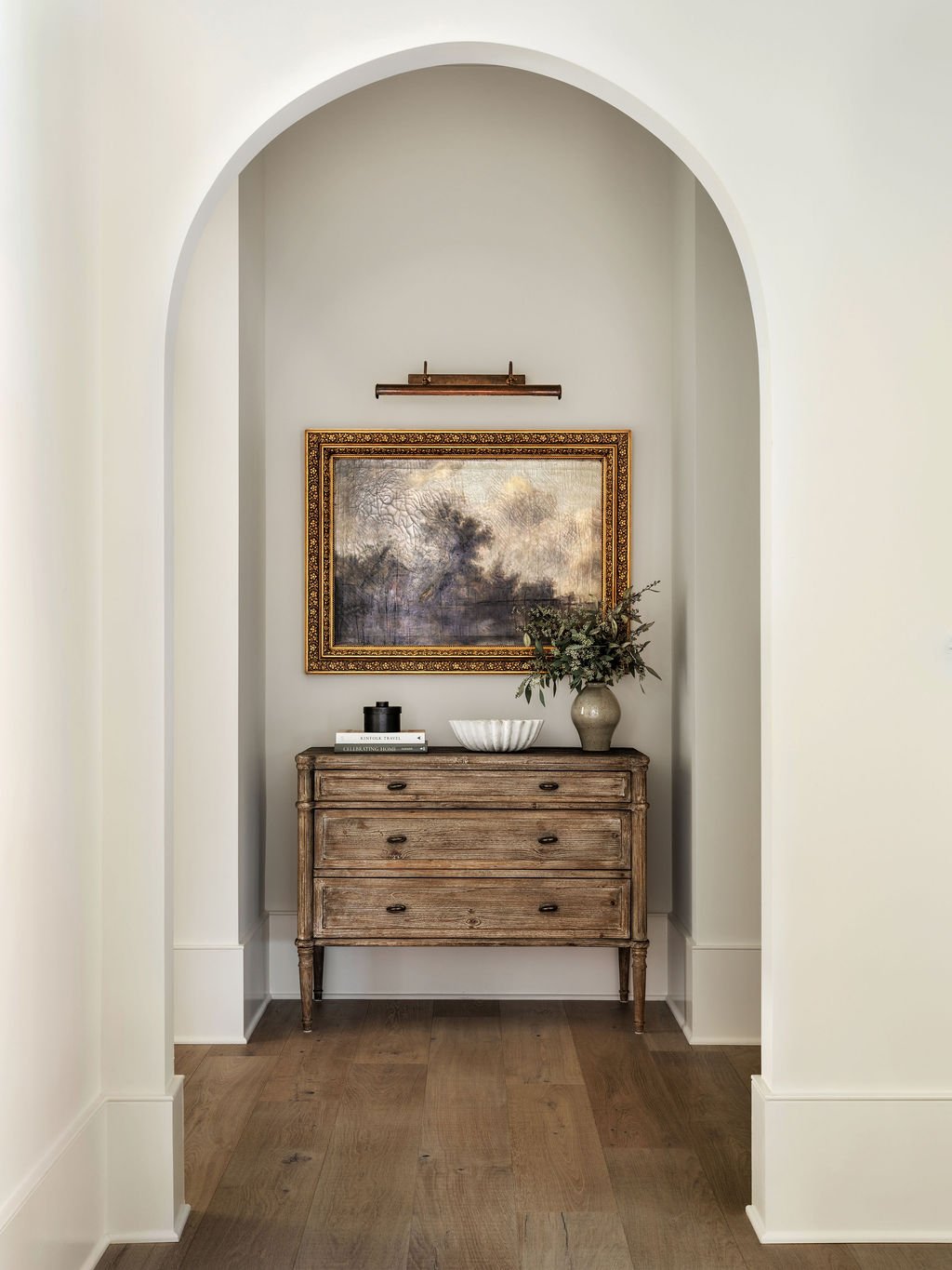 A wooden dresser with four drawers in an alcove, topped with a vase of greenery, a bowl, and a stack of books, featuring a framed landscape painting above.