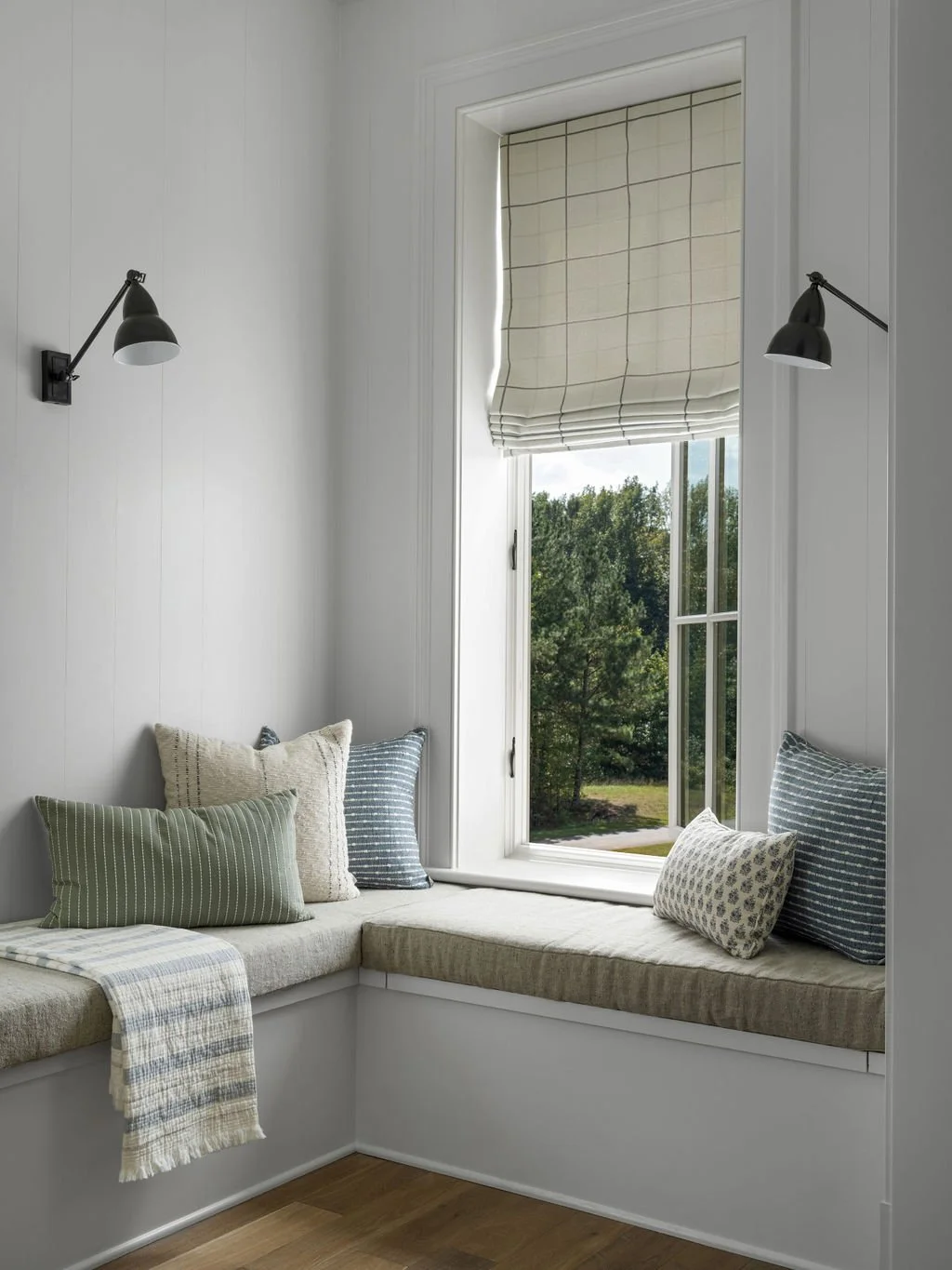 A cozy built-in window seat with multiple decorative pillows, beige cushion, accessed through a large window with a patterned Roman shade, in a bright white room with wall-mounted black lamps.