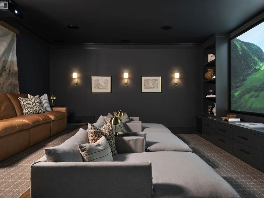 A modern home theater room with a large screen on the right, gray seating with pillows, and a dark wall with framed artwork and wall-mounted lamps.