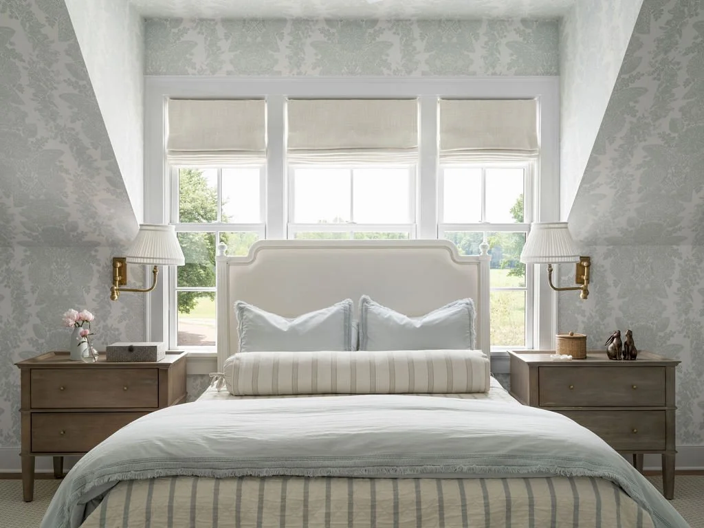 A neatly made bed with white pillows, a striped bolster pillow, and a bedspread with subtle stripes, flanked by two wooden nightstands with lamps and decor, and a large window with blinds behind the bed in a bright, cozy bedroom.