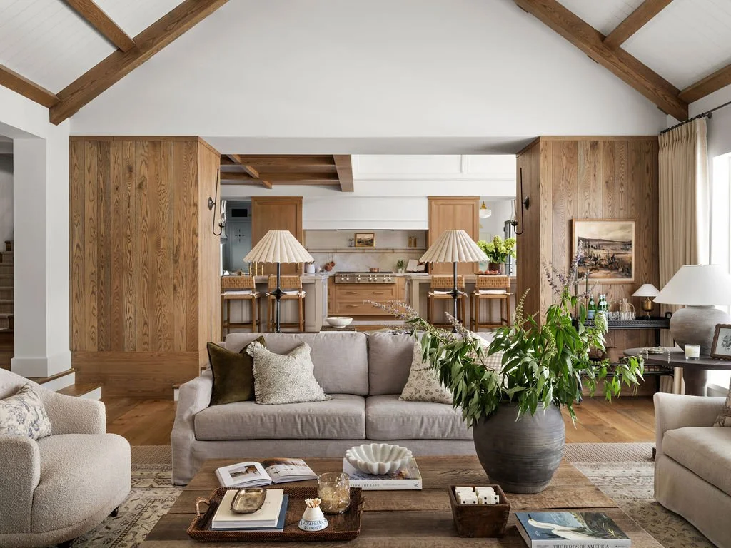 Living room with beige sofas, throw pillows, a large vase with green plants, a wooden coffee table with books and decorative items, wooden paneling, and a view into the kitchen with bar stools and pendant lamps.