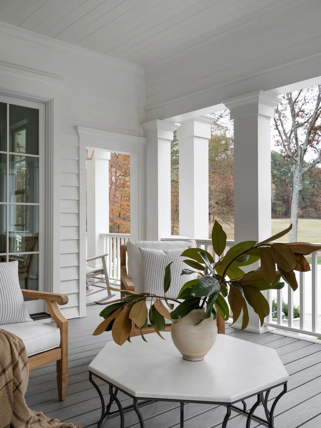A porch with white walls and columns, featuring wooden chairs with cushions, a white table with a potted plant, and trees visible in the background.