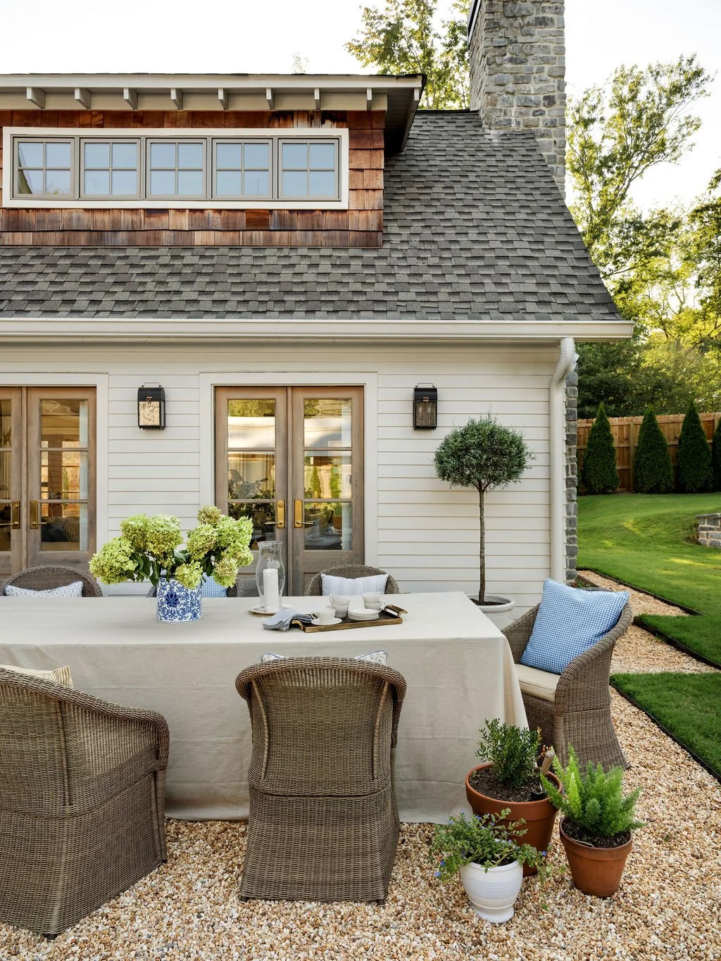 Outdoor dining area with a table and wicker chairs, potted plants, and a backyard with a house featuring a mix of siding, shingles, and stone, surrounded by trees and a lawn.